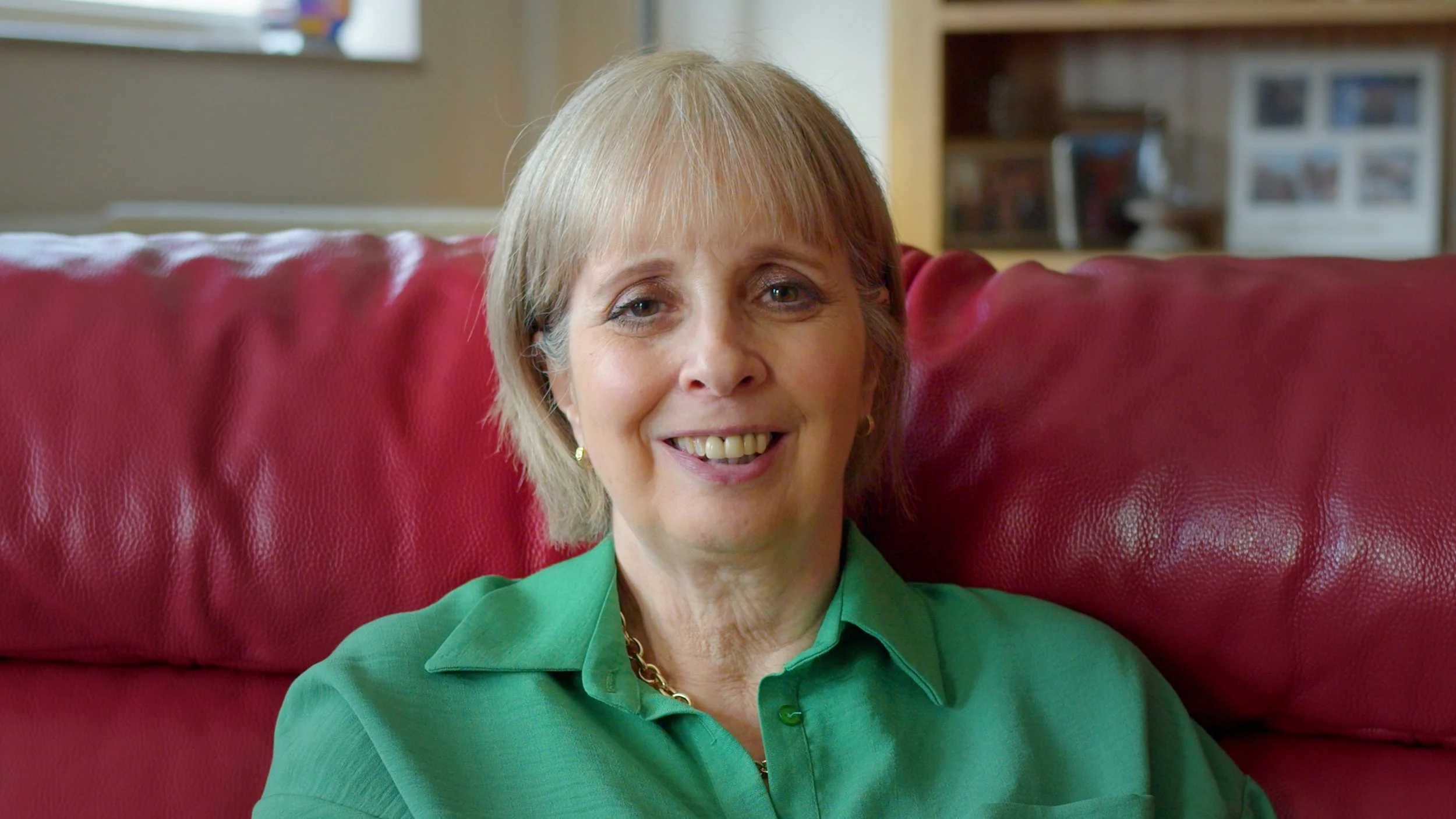 A woman with light brown hair, wearing a green shirt and gold jewelry, smiling while sitting on a red leather chair in a room with photographs on a wall in the background.