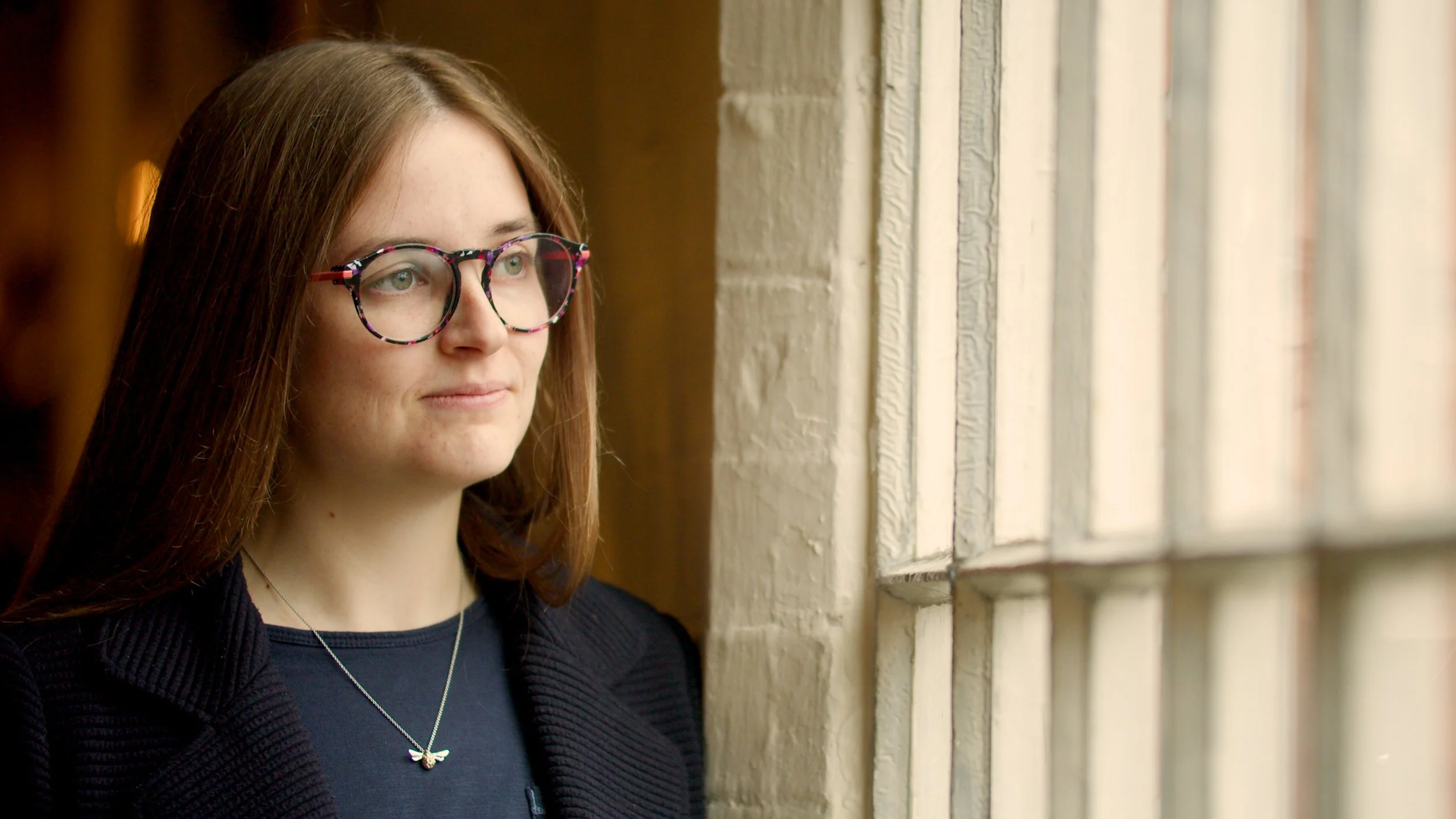 A young woman with long red hair and colorful glasses looking out a window with white brick framing.