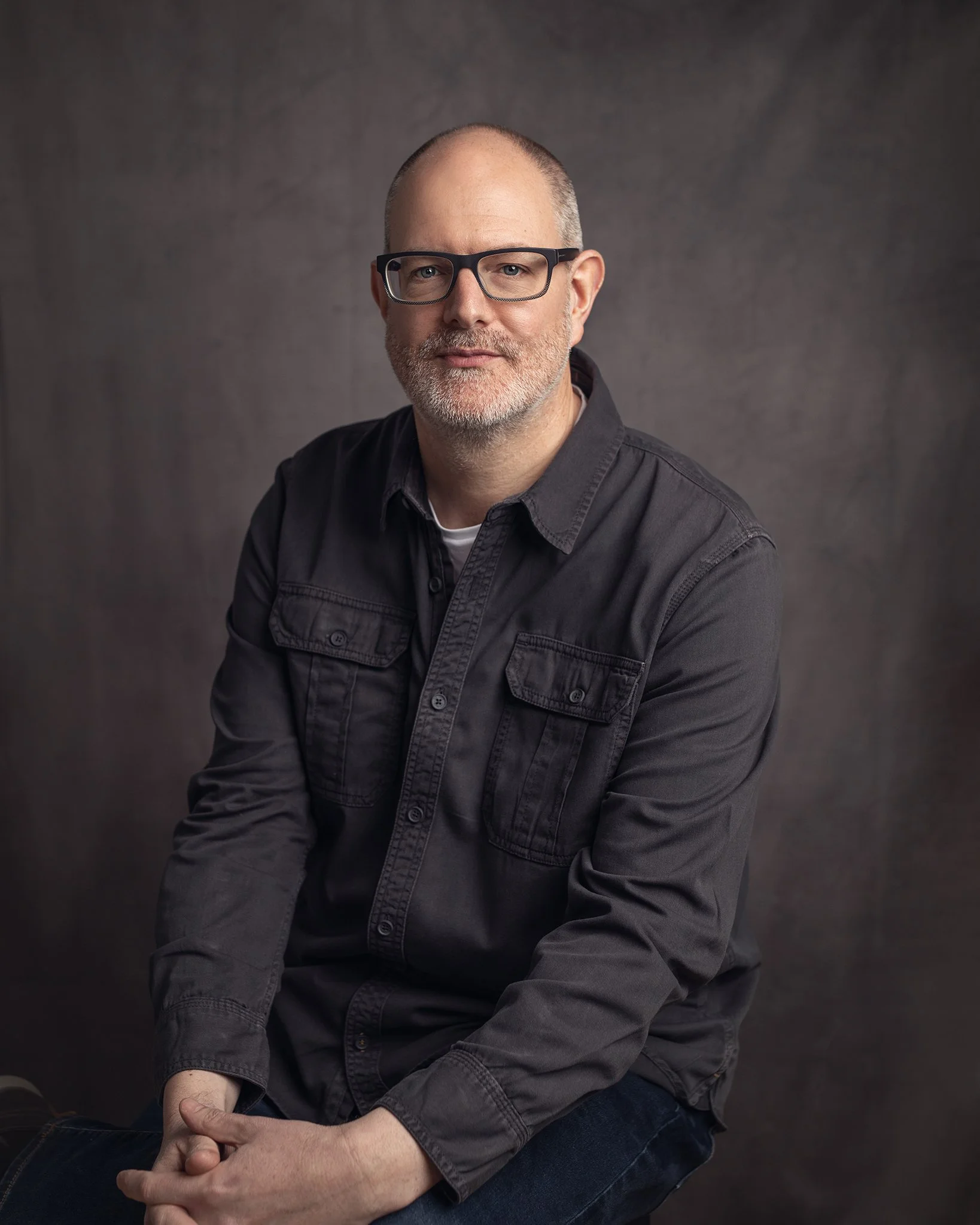 A middle-aged man with glasses, short beard, dressed in a dark shirt, sitting against a plain dark background.
