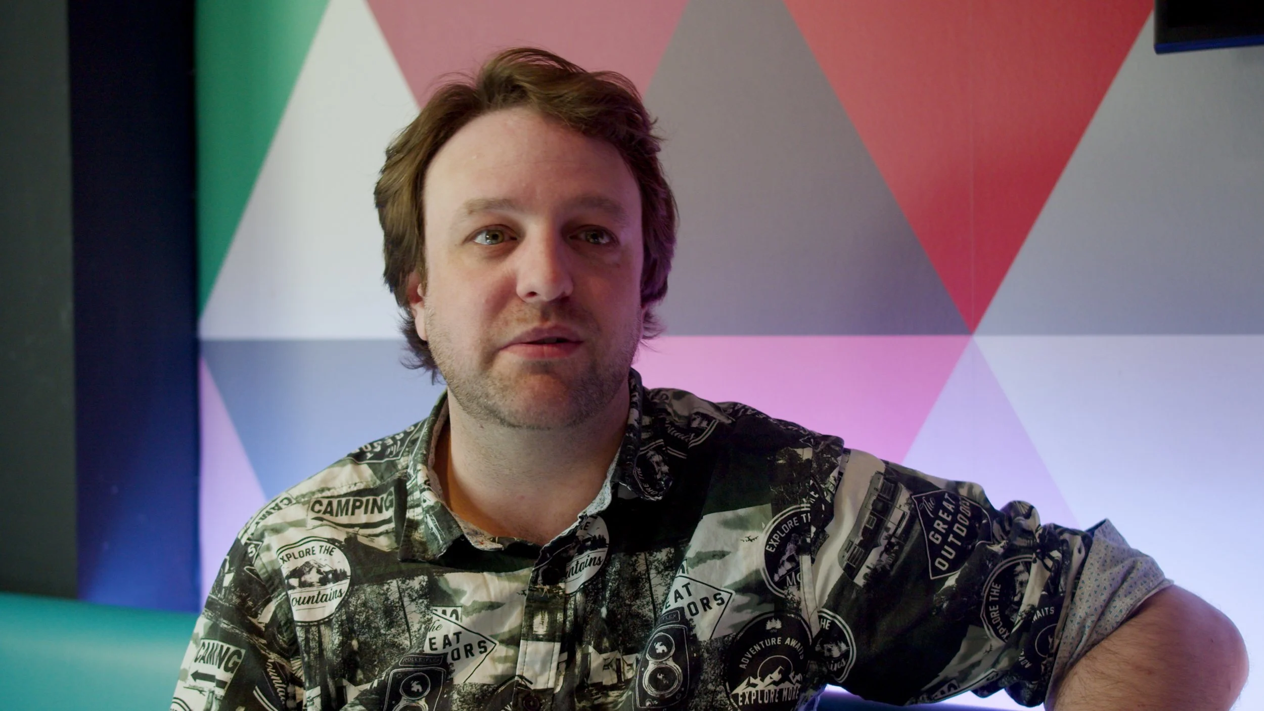A man with brown hair and a beard wearing a patterned shirt with outdoor and camping logos, sitting in front of a colorful, geometric background.