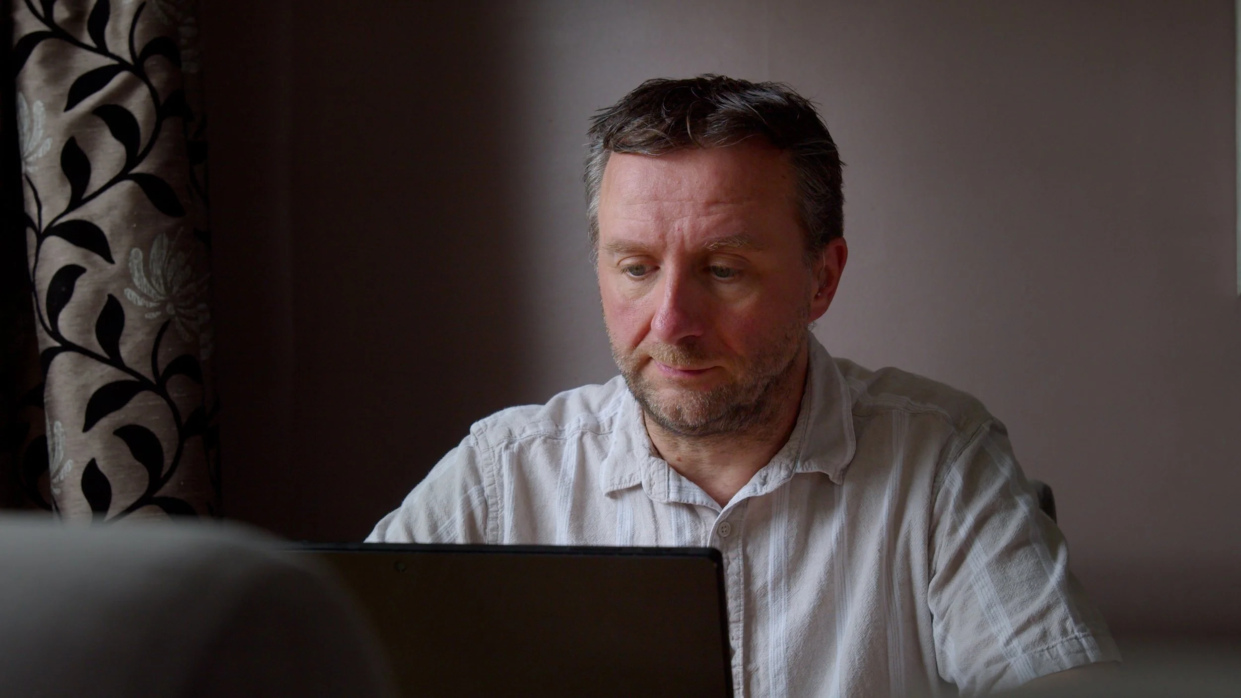 A man with short dark hair and a beard, wearing a light-colored, striped button-up shirt, looking down at a laptop in a room with dark walls and a patterned curtain on the left side.