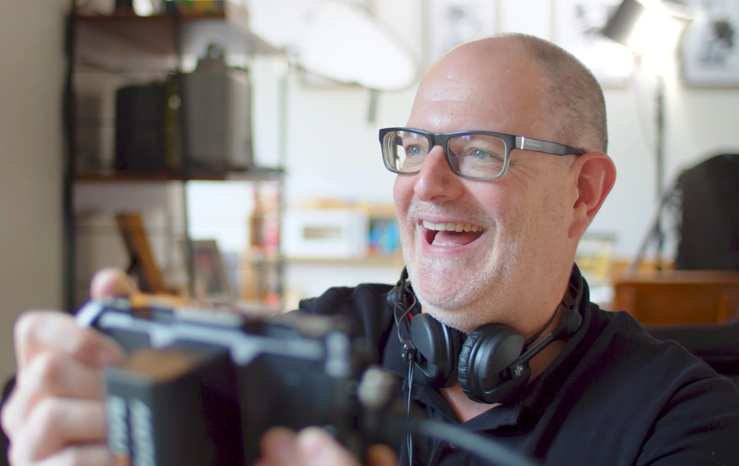 A middle-aged man with glasses and headphones around his neck is smiling and looking at his phone.