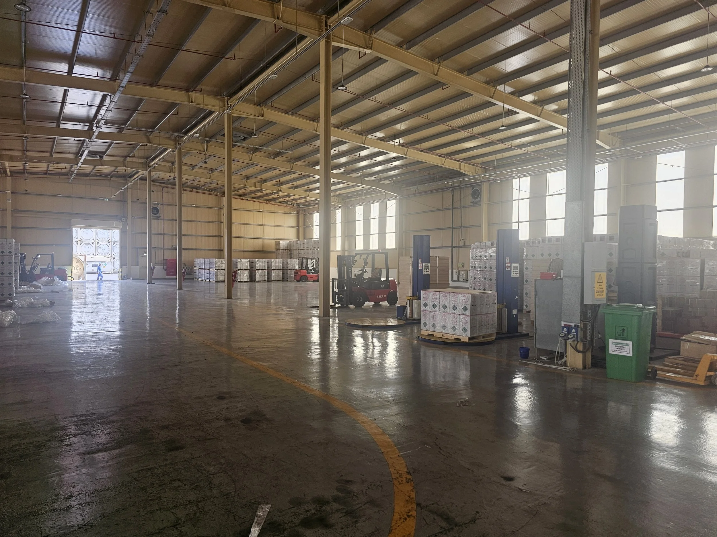Interior of a warehouse with a high ceiling, metal beams, and large windows letting in natural light. There are pallets of boxes, a green trash bin, and forklifts present.