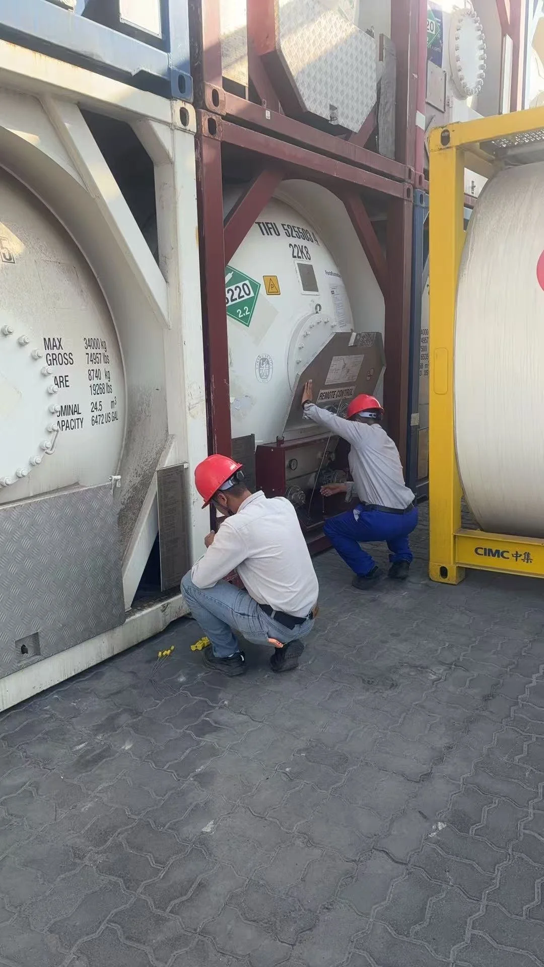 Two workers wearing safety helmets inspecting a large white tank container at an industrial site.