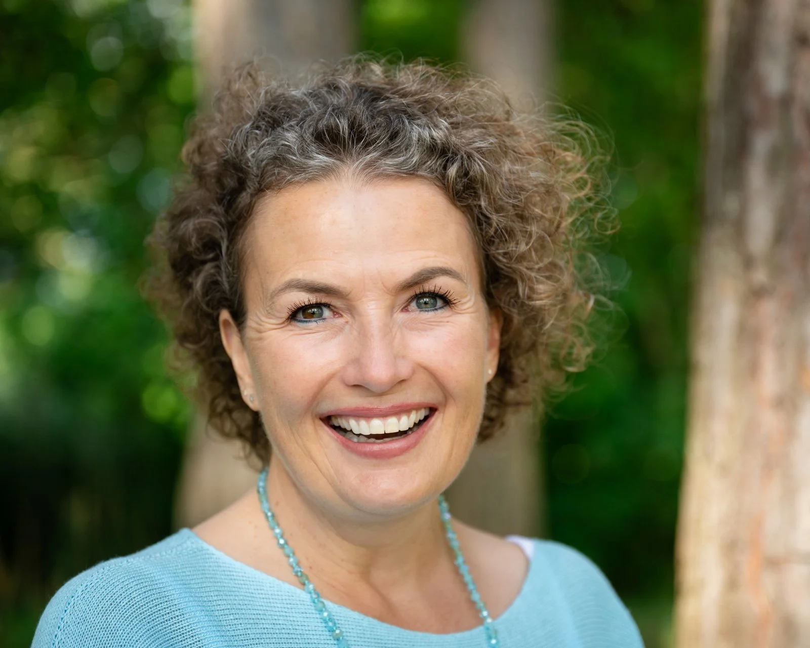 Lächelnde Frau, Barbara Wyser, mit hellblauen Augen, lockigem Haar, trägt einen hellblauen Pullover und eine turquoise Kette im Freien mit grüner, bunter Natur im Hintergrund.