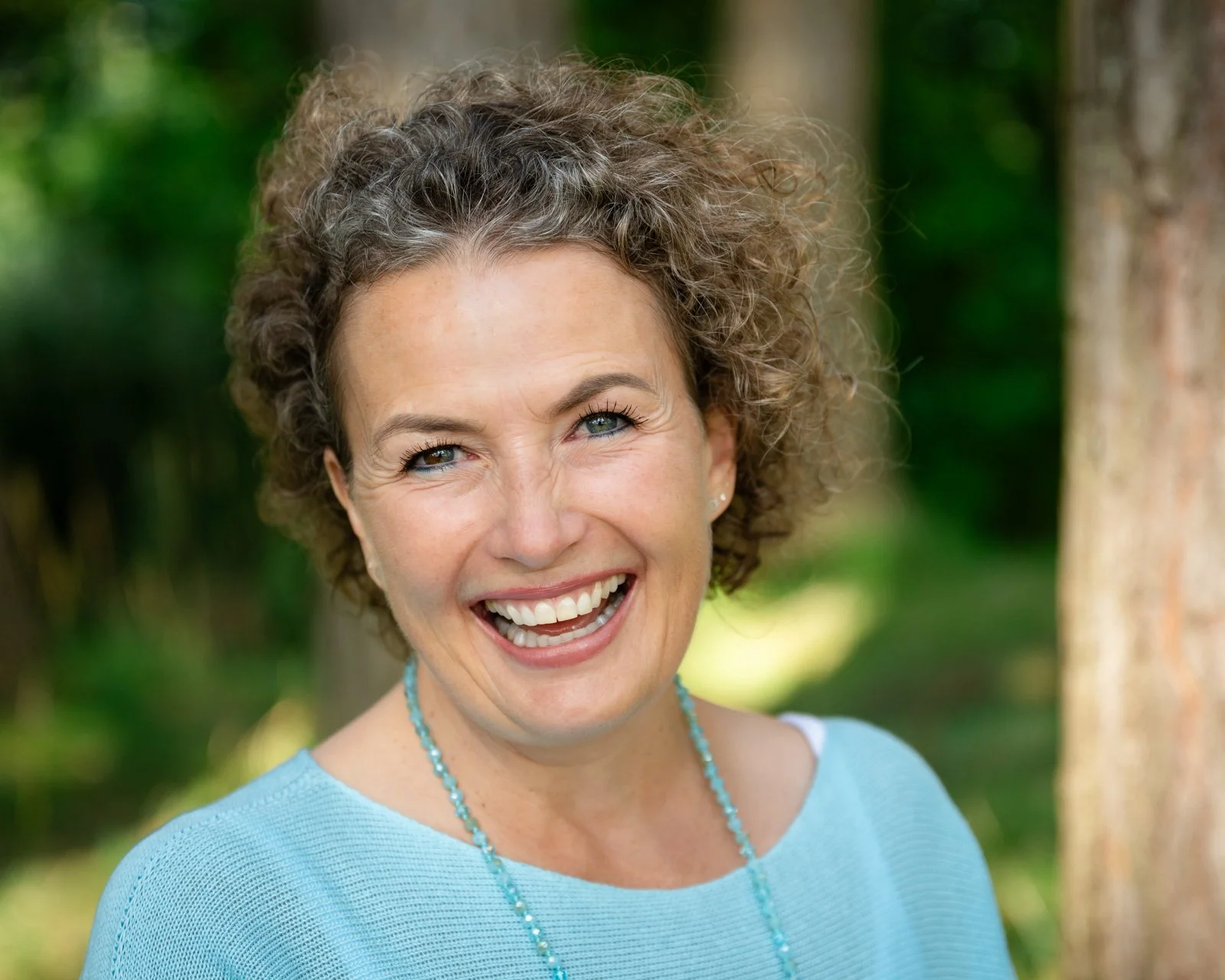 Lächelnde Frau, Barbara Wyser - im Freien mit kurzen, lockigen braunen Haaren, blauer Kleidung und Perlenkette, vor Baum im grünen Hintergrund.