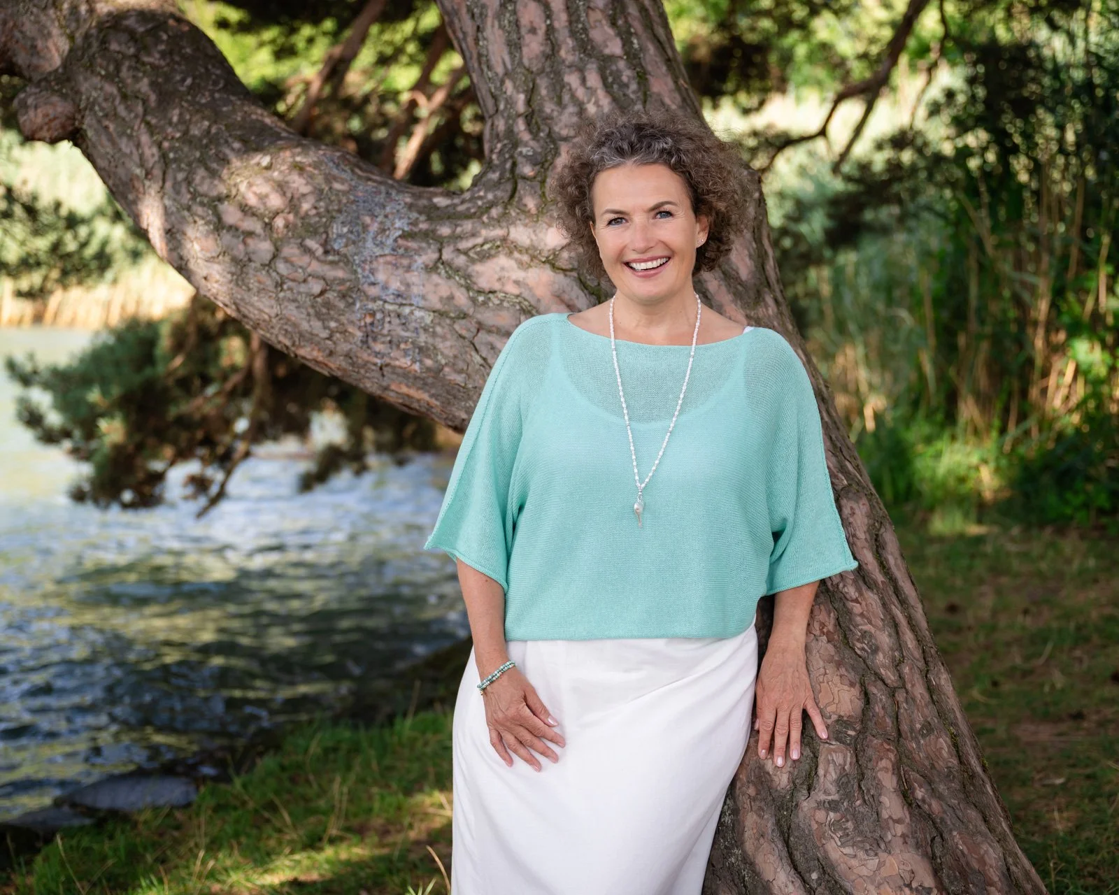 Eine lächelnde Frau - Barbara Wyser - mit kurzen, lockigen Haaren, in hellblauer Bluse und weißem Rock, lehnt an einem Baum neben einem Fluss im Freien bei Sonnenlicht.