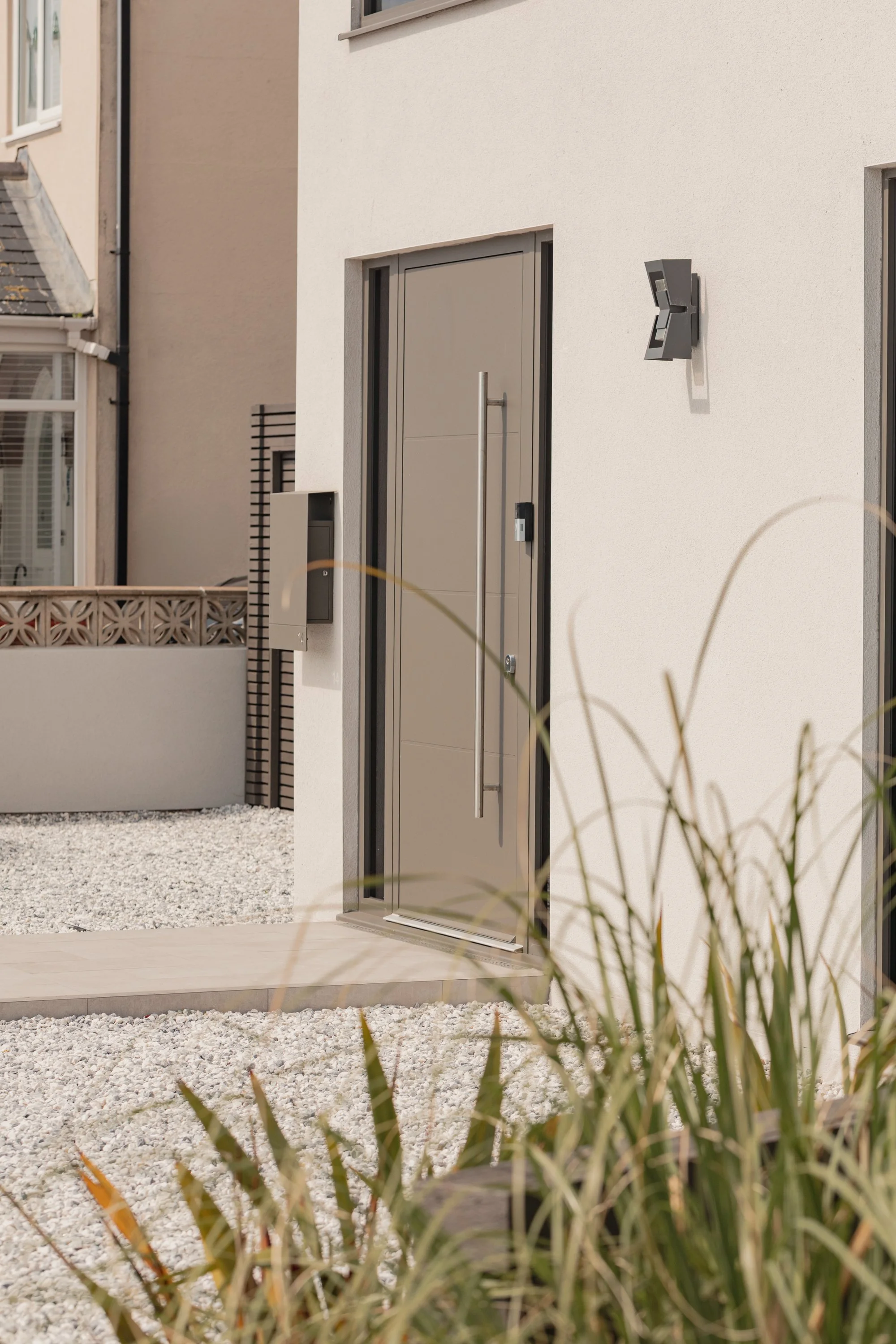 Modern front entrance of a house with a gray door, a long vertical handle, and a keypad lock. There is a wall-mounted light fixture and an intercom on the white exterior wall. The area is landscaped with white gravel and some plants in the foreground.