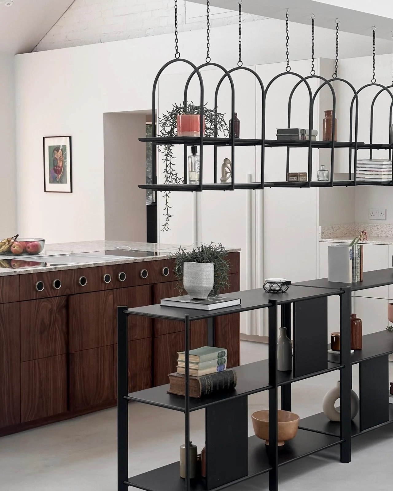Modern kitchen interior with black metal shelving and a wooden cabinetry with a marble countertop, decorated with potted plants, books, and decorative objects.