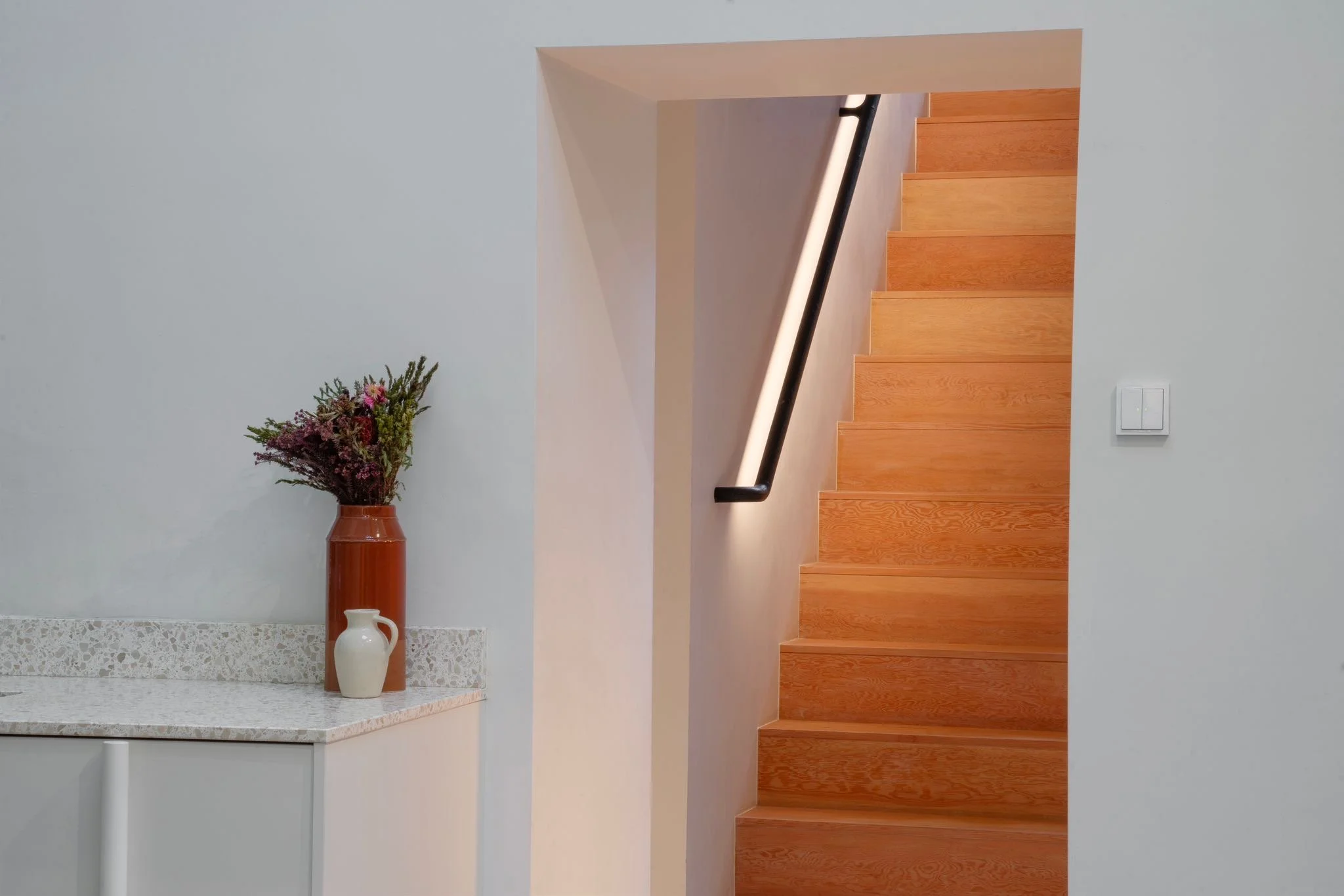 Interior view of a modern house showing a staircase with wooden steps and a black handrail, adjacent to a white wall with a light switch. To the left, a marble-topped cabinet holds two vases, one with dried flowers and the other empty.