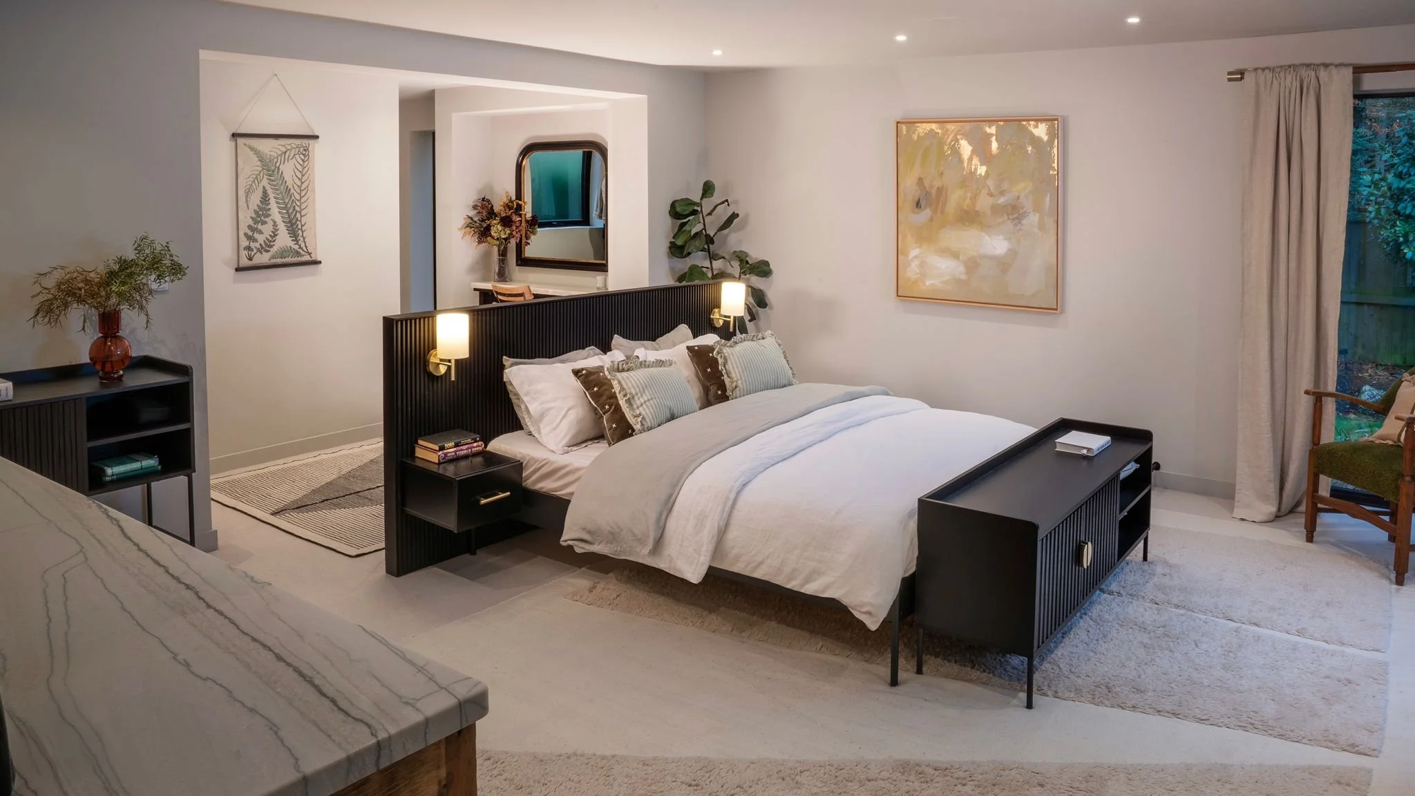 A cozy bedroom with a large bed that has white and striped pillows, a black headboard, and nightstands with lamps. There's a chair near a window, curtains, wall art, and decorative plants, creating a warm and modern ambiance.