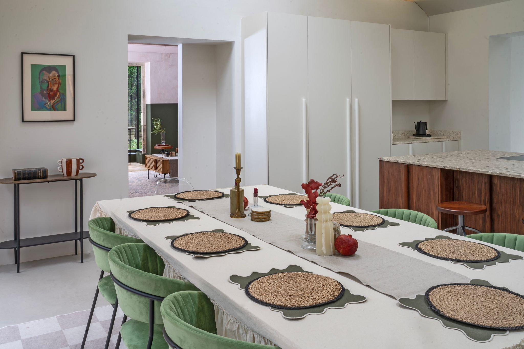 Dining room with a long table set with seven placemats, a white tablecloth, and various decorative items. There are green velvet chairs around the table. In the background, a kitchen with white cabinets and a marble countertop is visible, along with an open doorway leading to a room with large windows and patio doors.