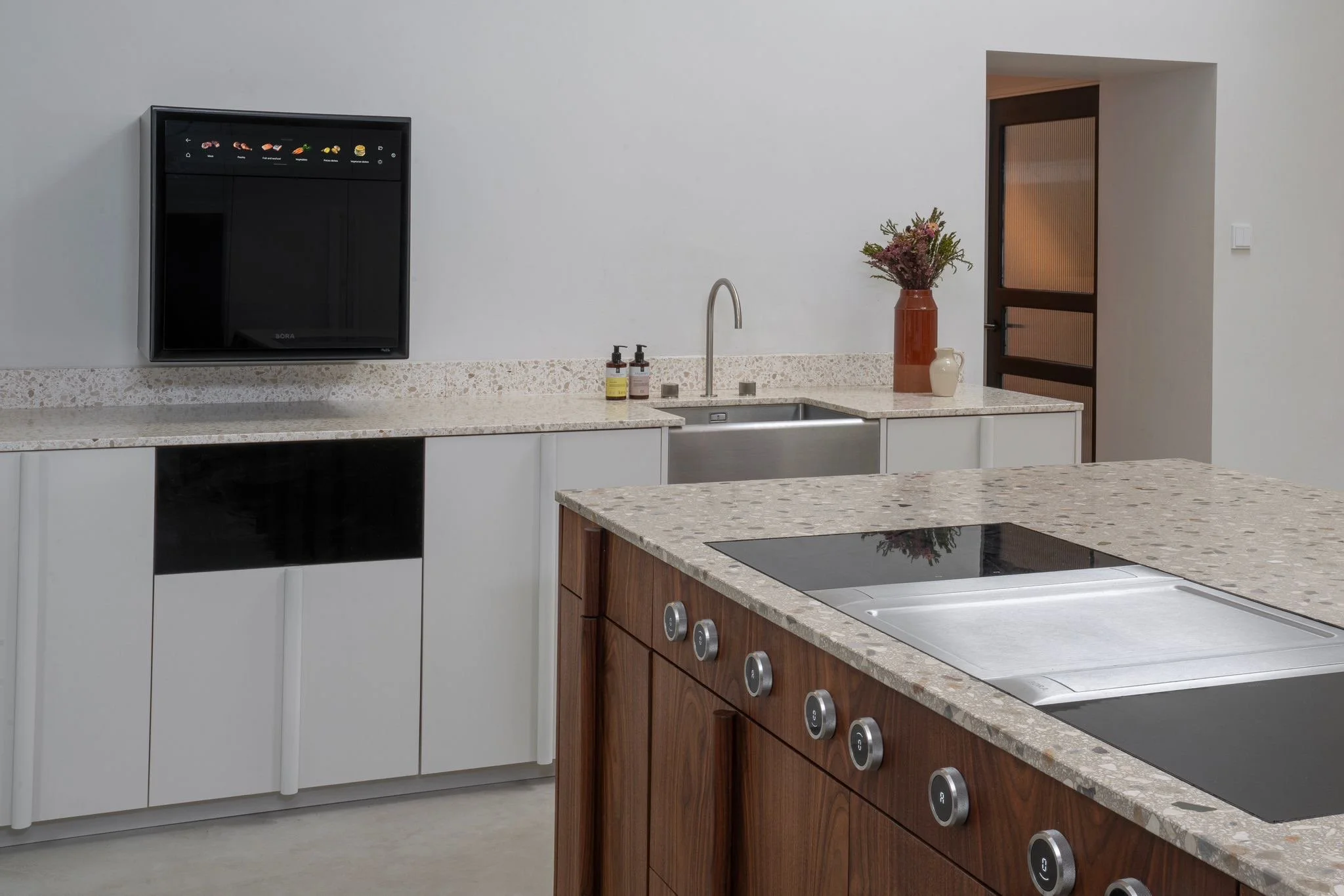 Modern kitchen with white and wooden cabinets, a built-in stove on a speckled countertop, a sink with a faucet, and a wall-mounted flat-screen TV. A door with glass panels is in the background, decorated with vases and flowers.