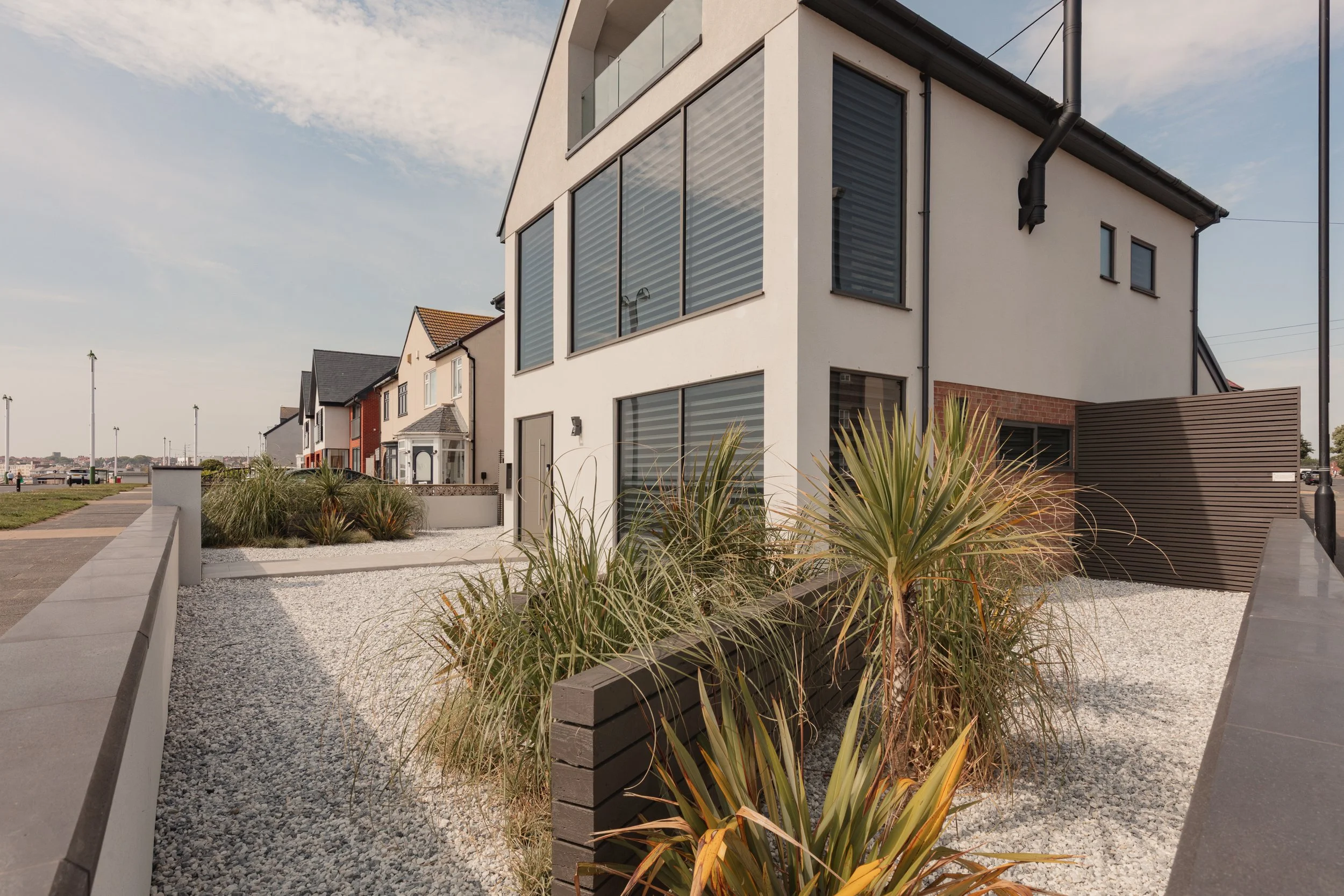 Modern multi-story house with large windows, landscaped yard with ornamental grasses, white gravel ground cover, and a brown wooden privacy screen.