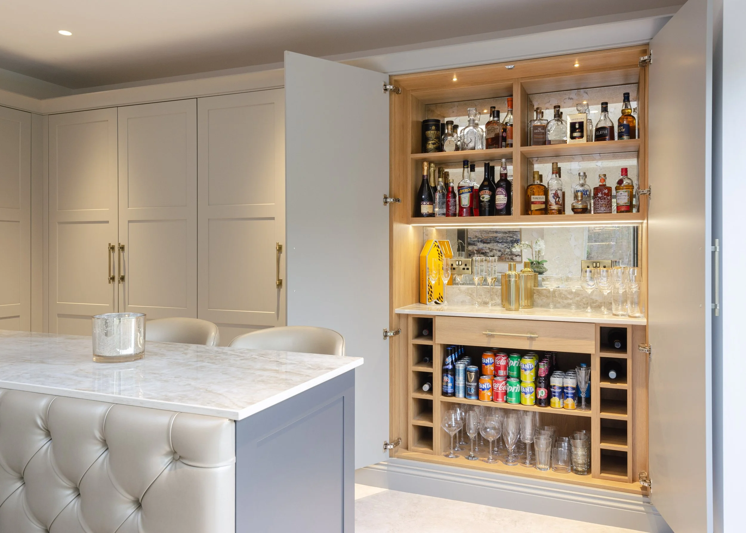 A built-in bar cabinet with glassware, liquor bottles, and soda cans inside a modern open kitchen.