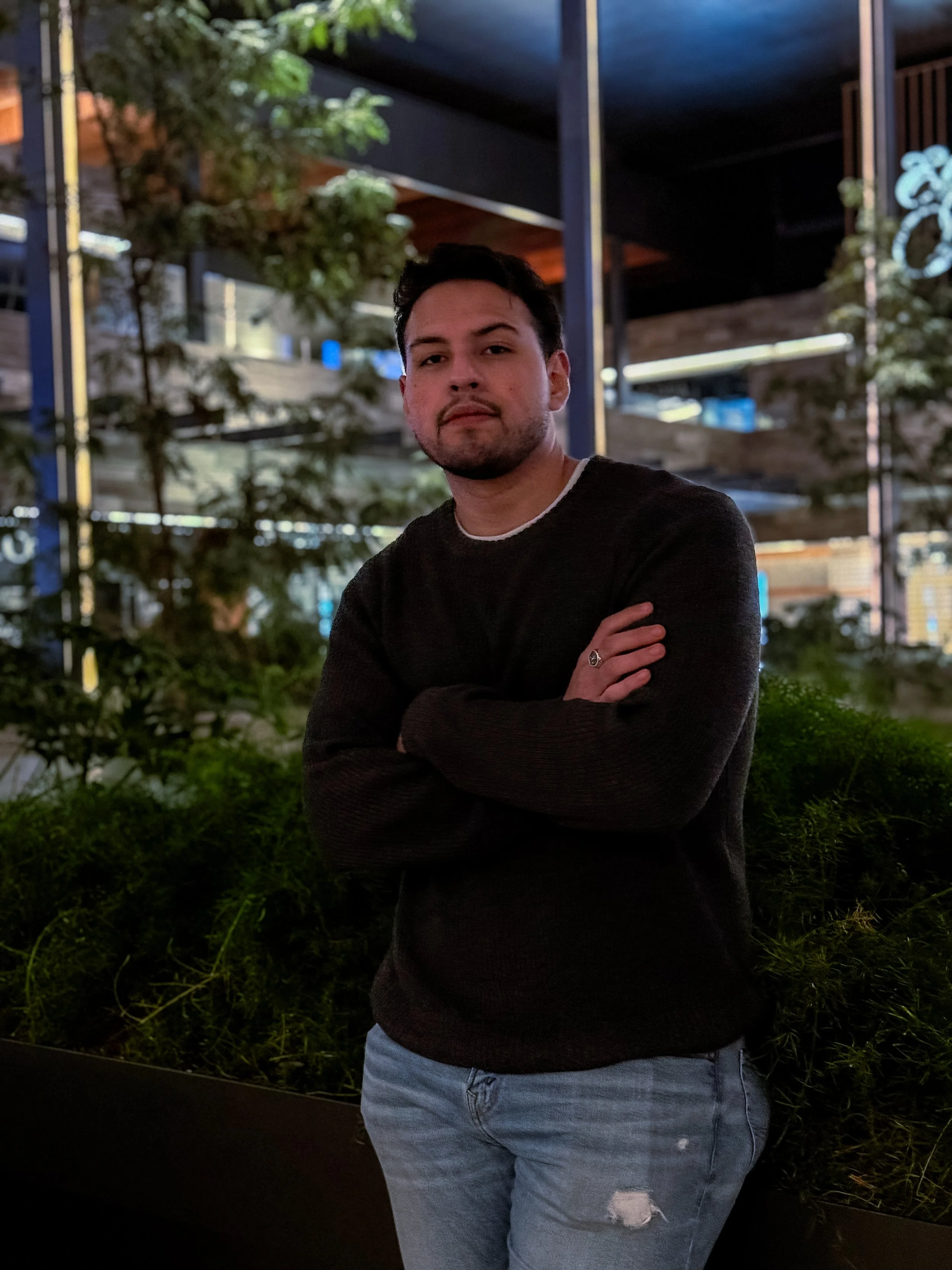 Joven de cabello oscuro y barba en un entorno nocturno con plantas y estructura metálica en el fondo, vistiendo suéter negro y jeans claros.