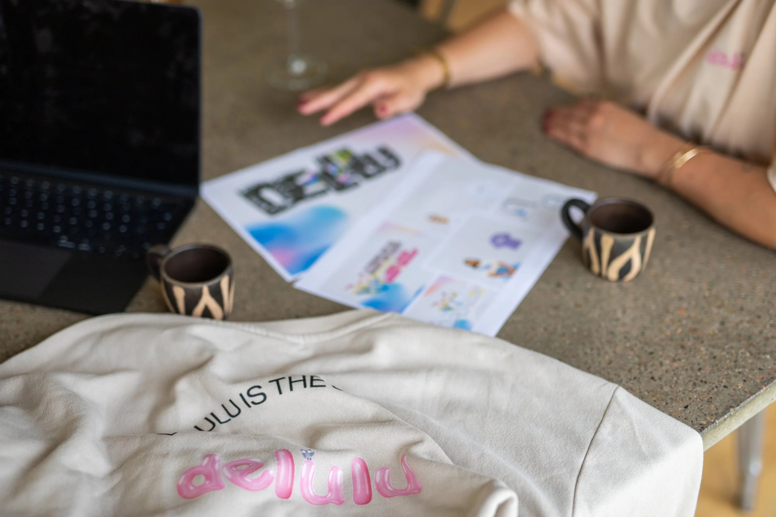 A person's arm is resting on a table with a laptop, two zebra-patterned mugs, and printed papers. A beige cloth with pink and black text is draped over the edge of the table.