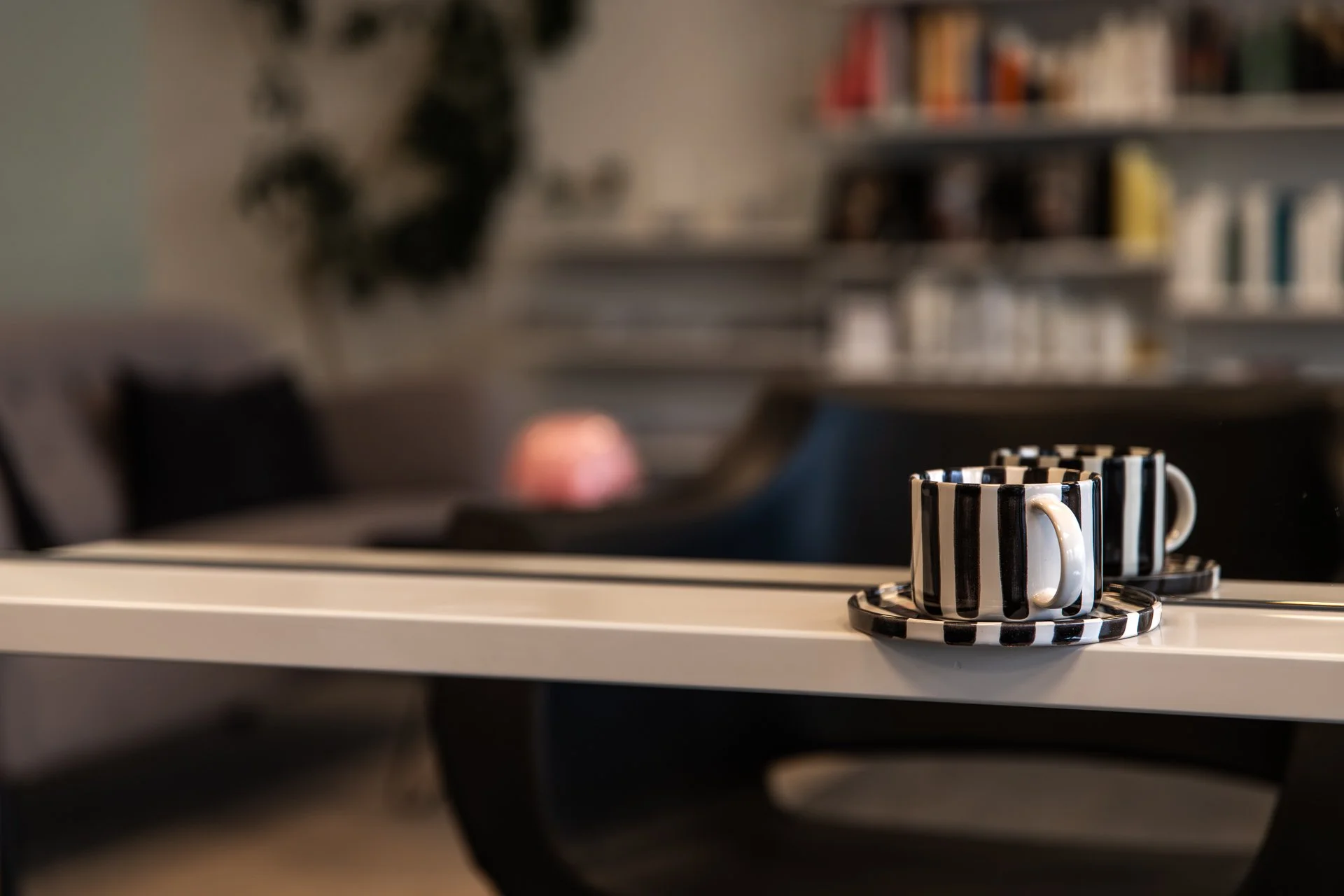 Two black and white striped ceramic coffee cups with matching saucers on a white surface, with a blurred background of a bookshelf and a chair.