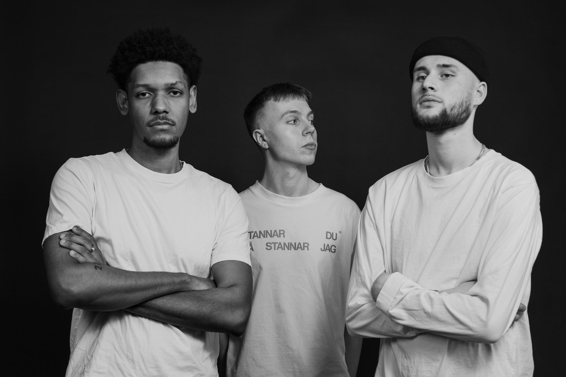 Black-and-white photo of three young men standing with arms crossed against a black background.