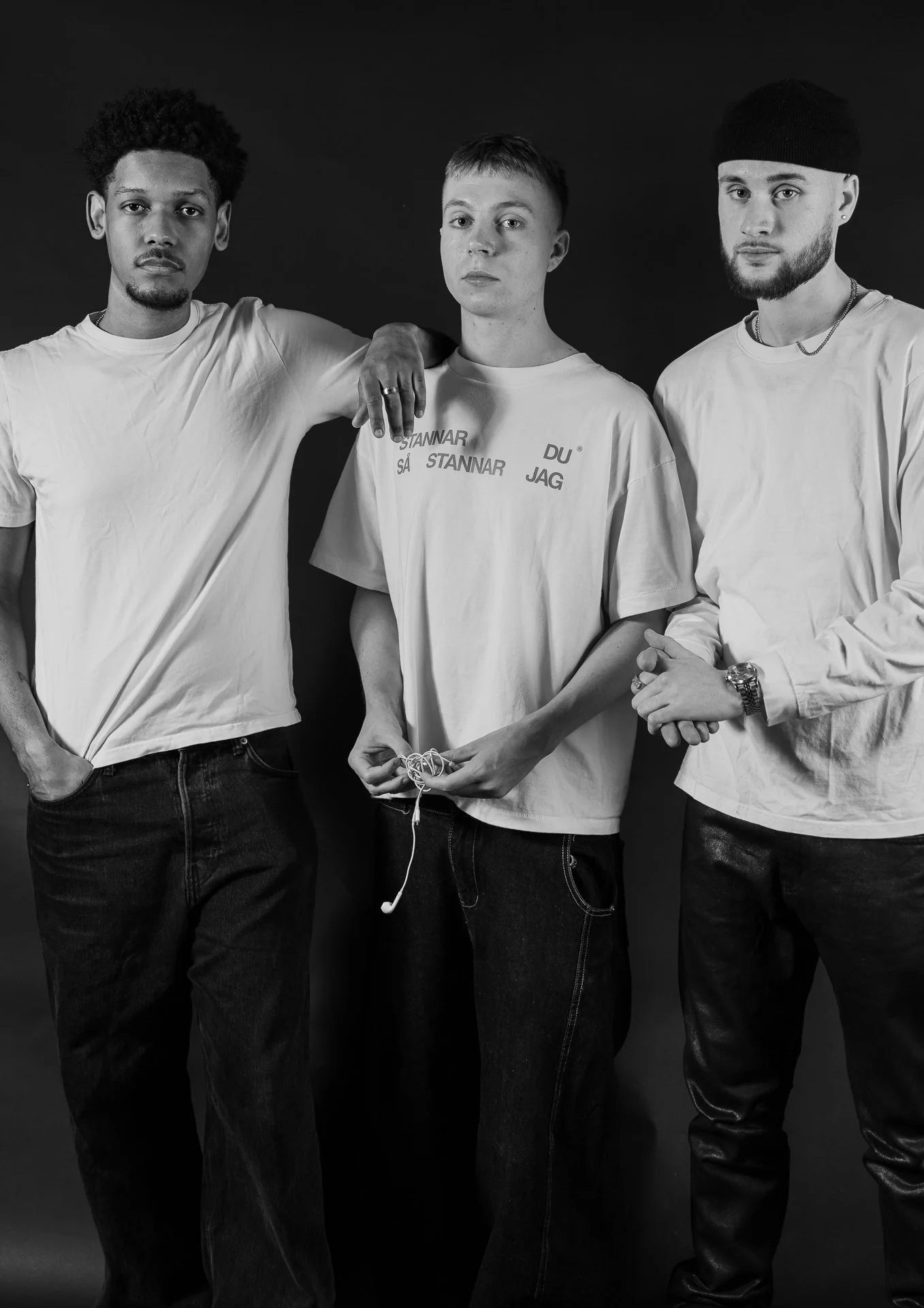 Three young men standing side by side in front of a black background, posing for a black and white photo, wearing casual clothes, with the middle man holding a pair of earphones.