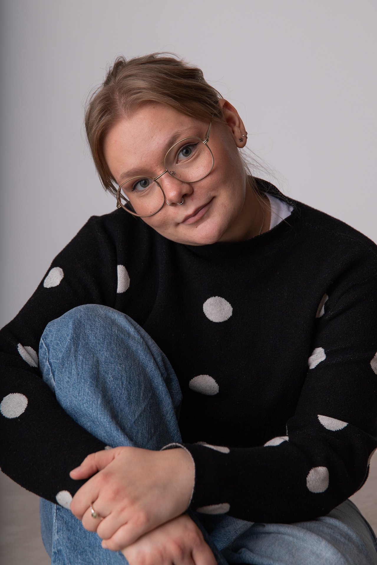 A young woman with light skin and short brown hair, wearing glasses and a black sweater with white polka dots, sitting with her arms crossed and smiling in front of a plain gray background.