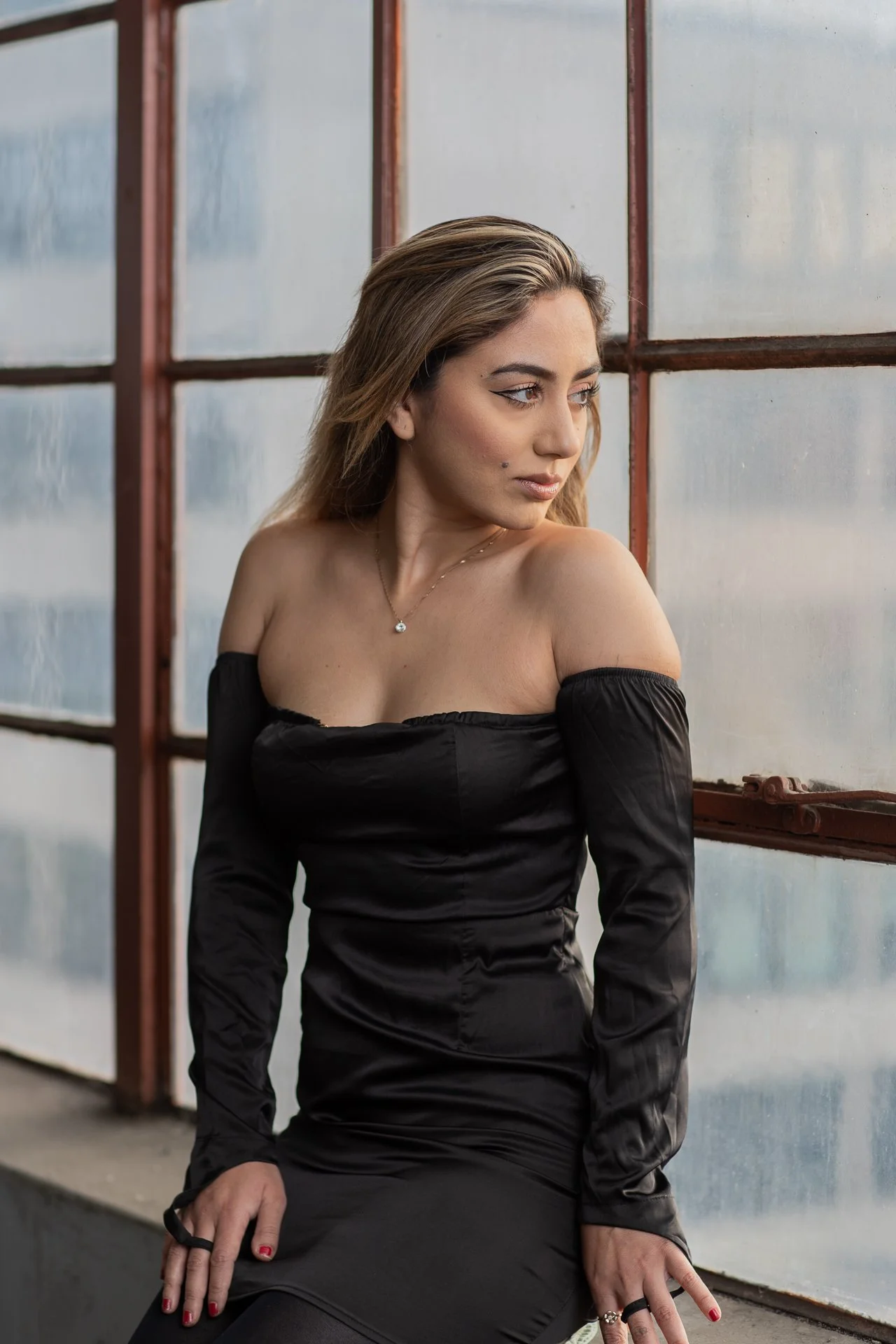 A woman with light brown hair, wearing a black off-the-shoulder dress, sitting near a large window with a reddish-brown frame, looking to the side.