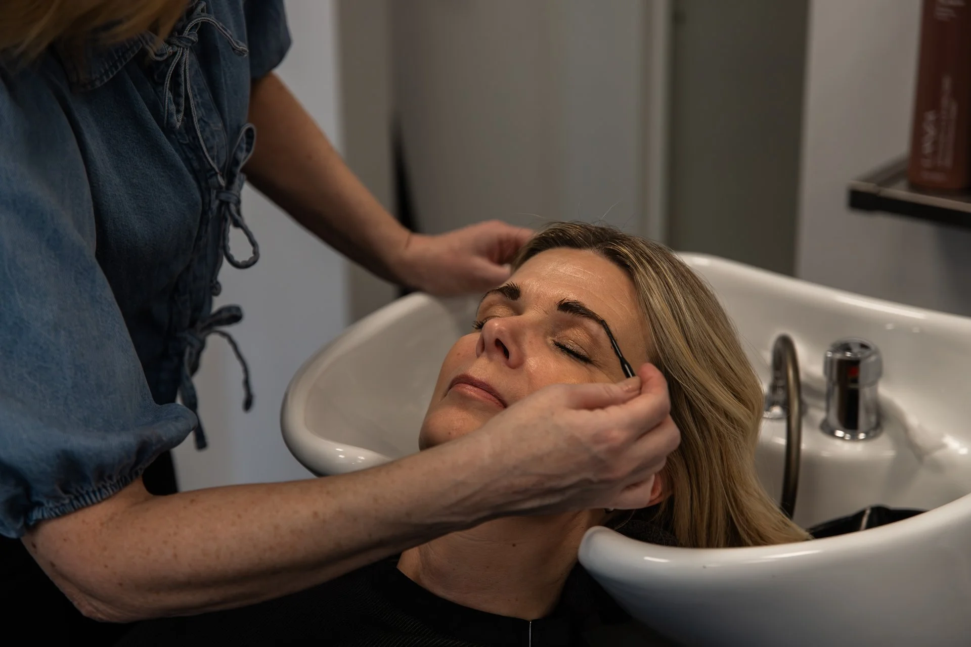 A woman is lying back in a salon sink with her head tilted, as a stylist applies makeup or grooming cosmetics near her eyebrows.