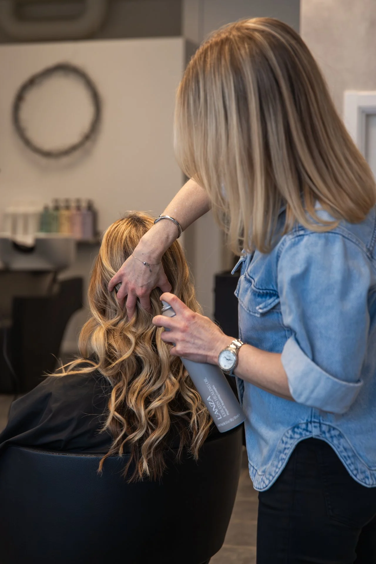A hairstylist spraying hair with a spray bottle while working on a client with long, wavy blonde hair sitting in a salon chair.
