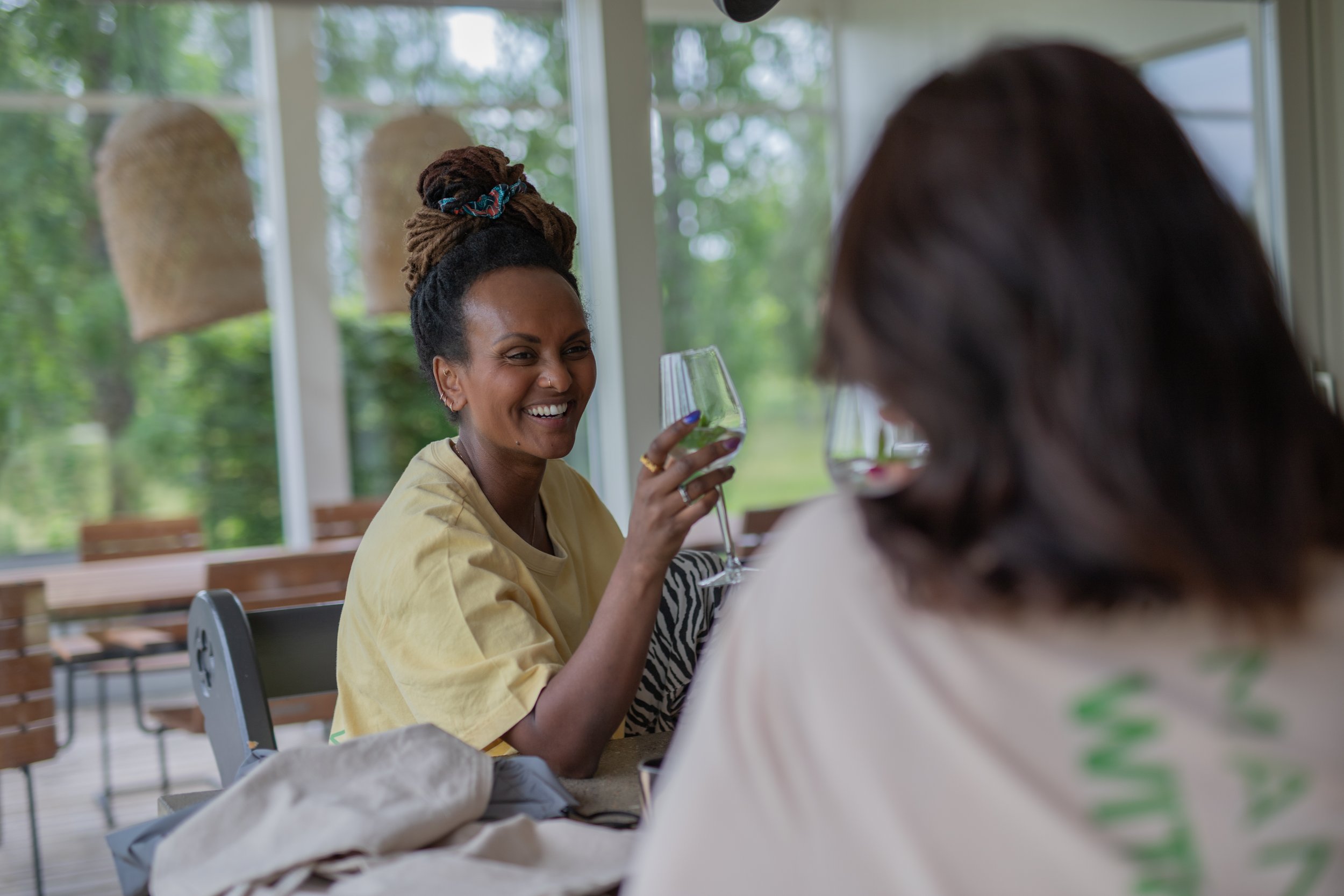 A woman with dreadlocks in a bun, wearing a yellow shirt, smiling and holding a glass of wine at a table indoors near large windows with a view of greenery.
