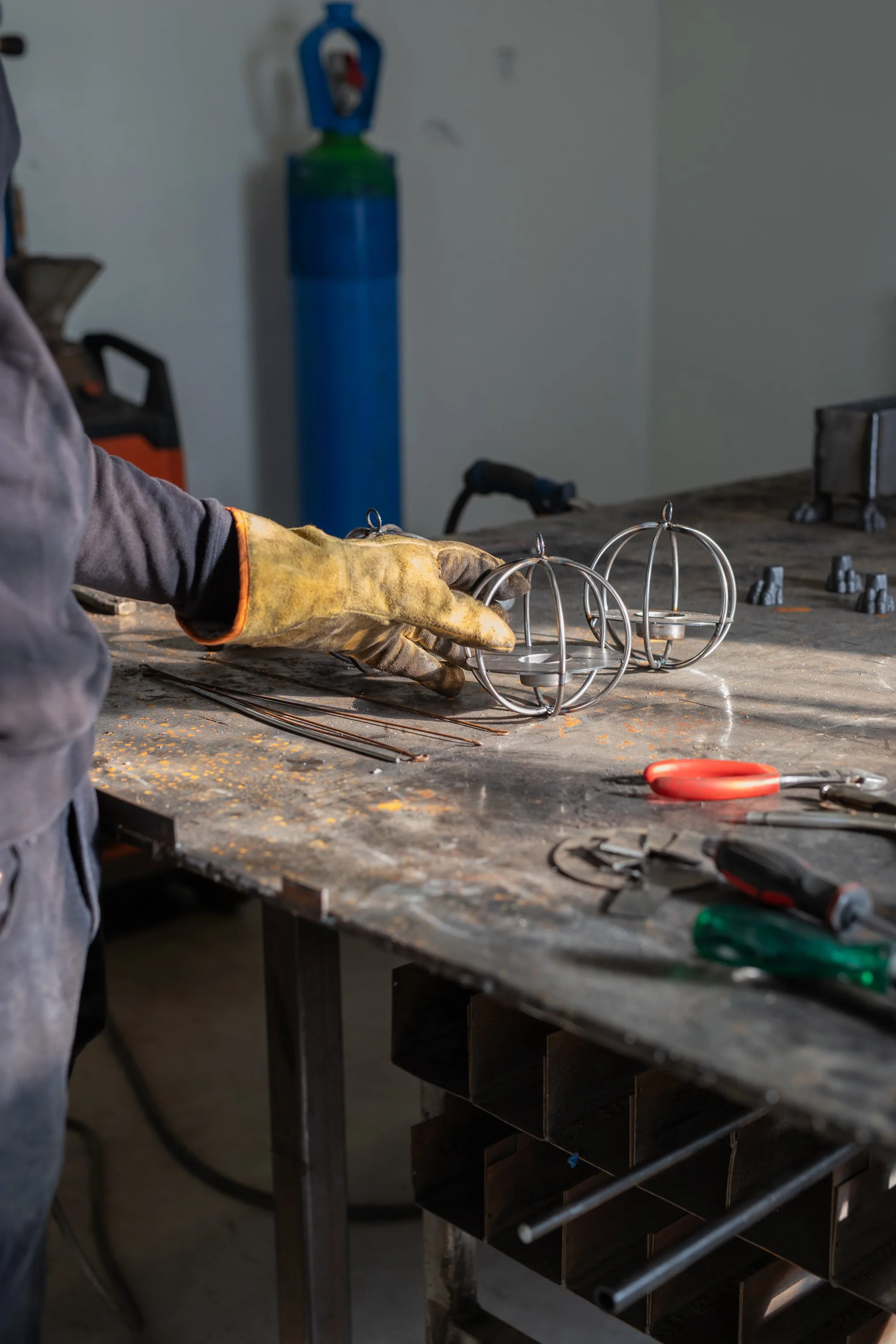 A worker's gloved hand working on metal spherical wire frames with other tools and materials on a workshop table.
