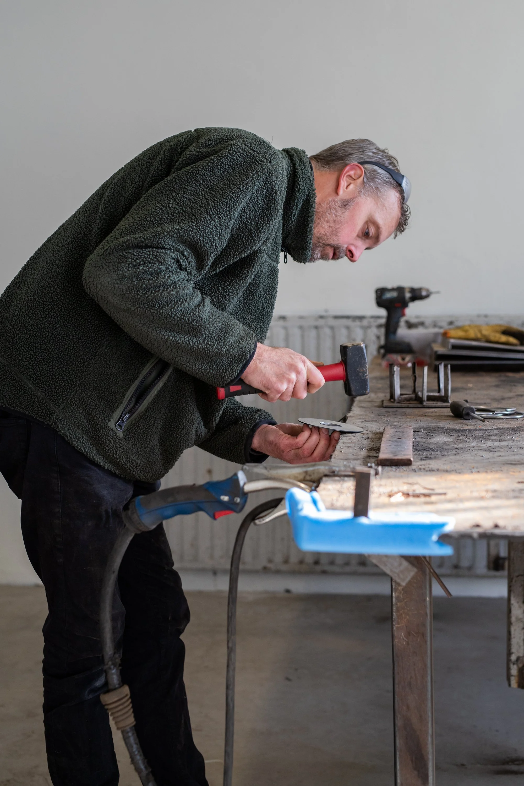 A man working in a workshop, hammering a piece of metal on a wooden workbench with various tools and equipment around.