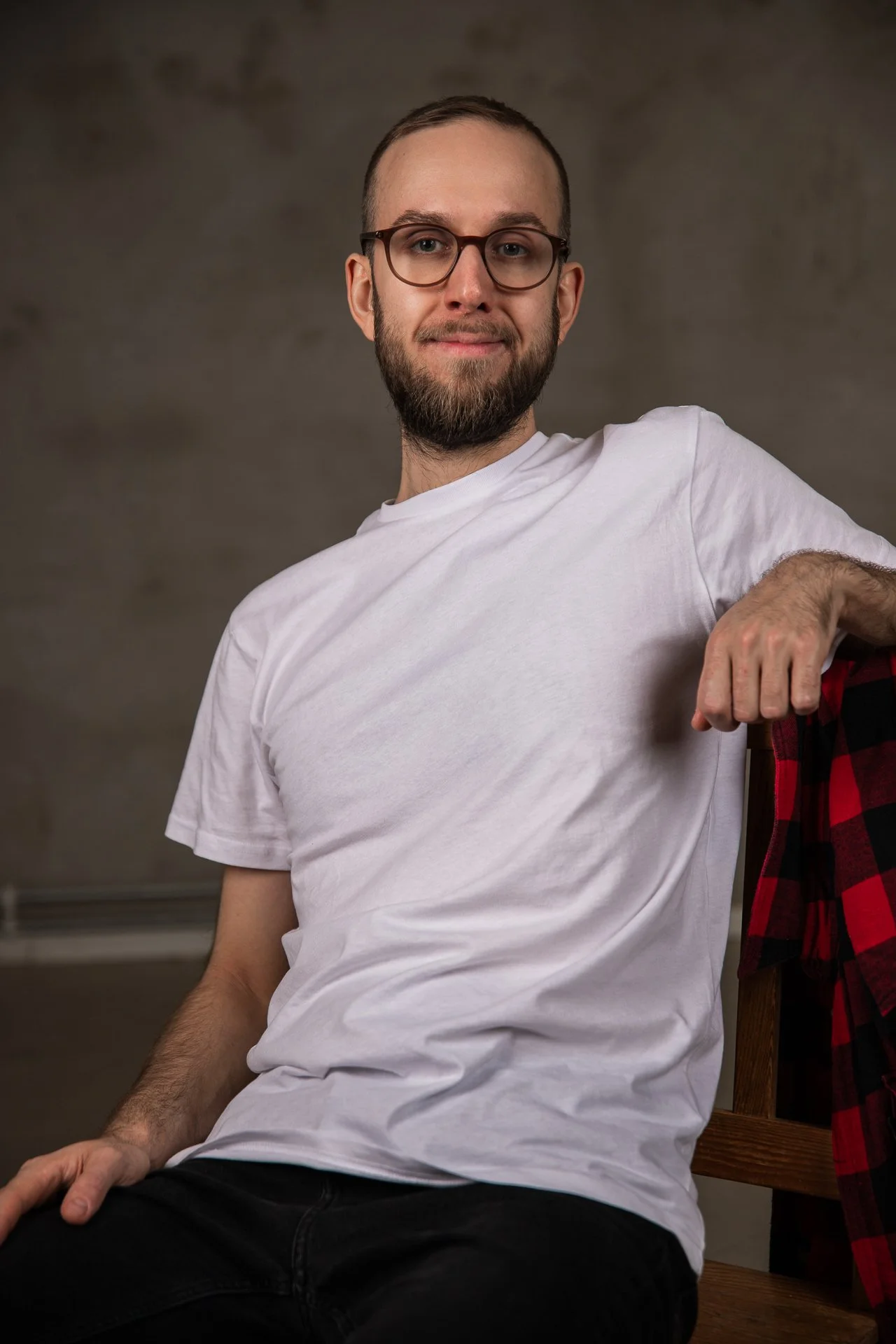 A man with glasses and a beard wearing a white T-shirt, sitting on a wooden chair with a red and black checkered shirt draped over the back.