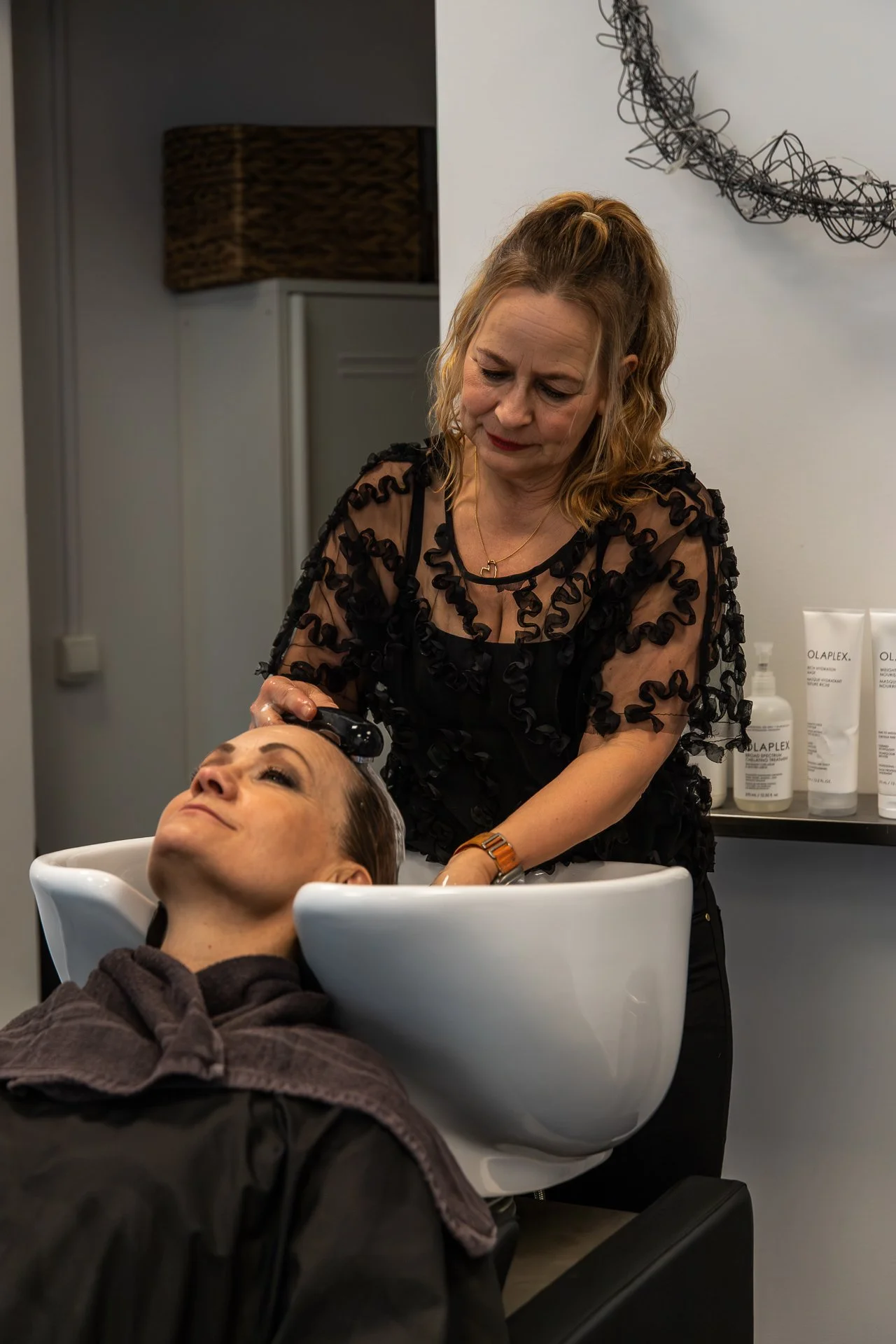 A woman with curly blonde hair washing a client's hair in a salon, with hair care products on a shelf behind her.