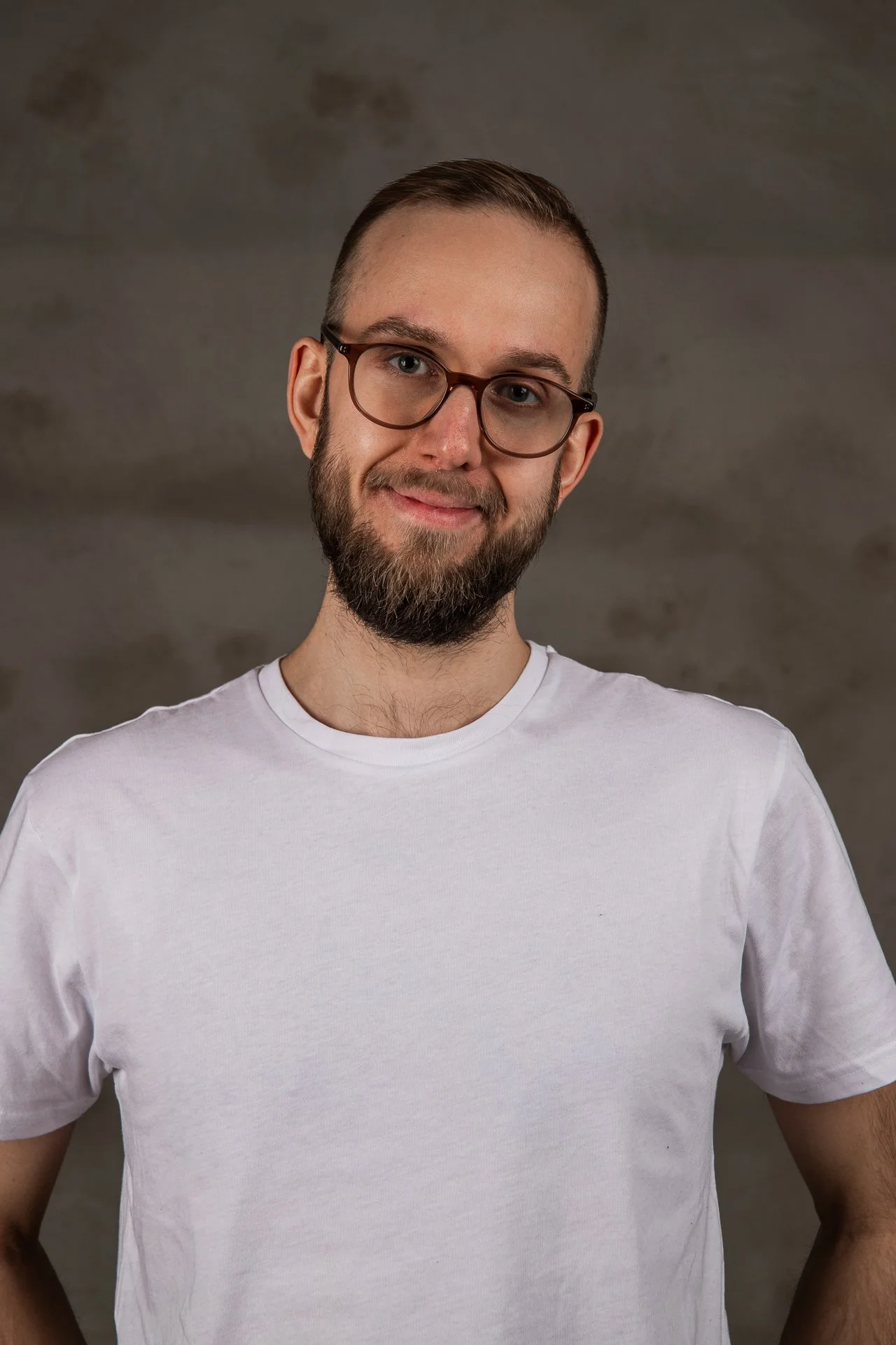 A young man with glasses, a beard, and a white t-shirt standing against a neutral background.