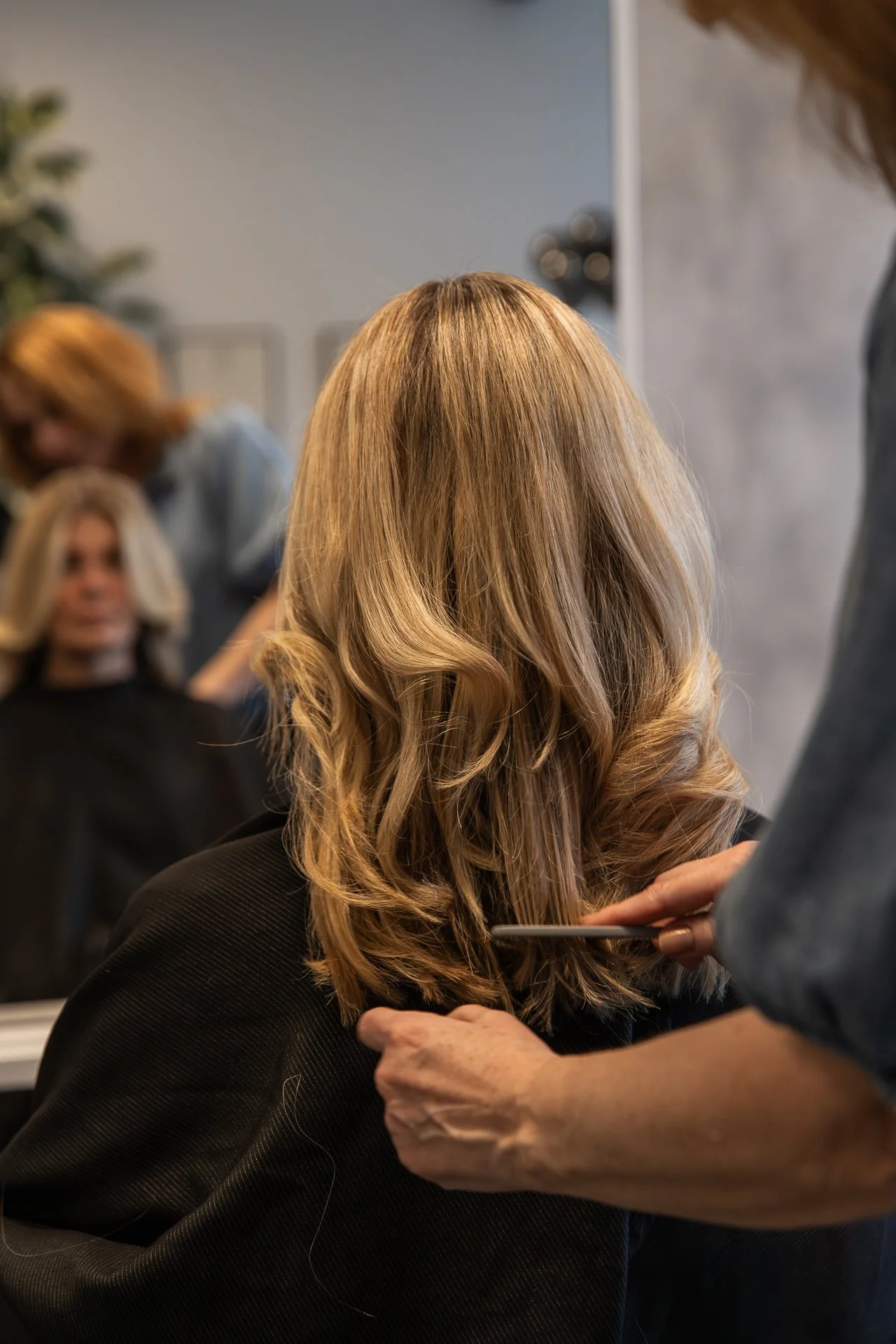 A person with long, wavy blonde hair sitting in a salon chair while a stylist works on their hair, with a mirror and another hairstylist in the background.