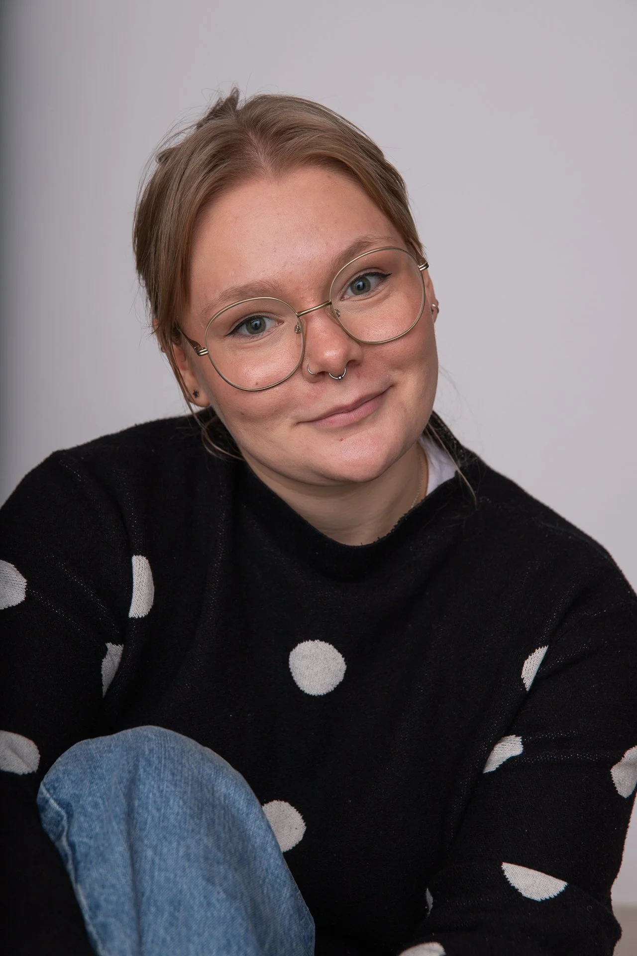 A young woman with short red hair, round glasses, and a septum piercing, smiling slightly while wearing a black sweater with white polka dots and blue jeans.
