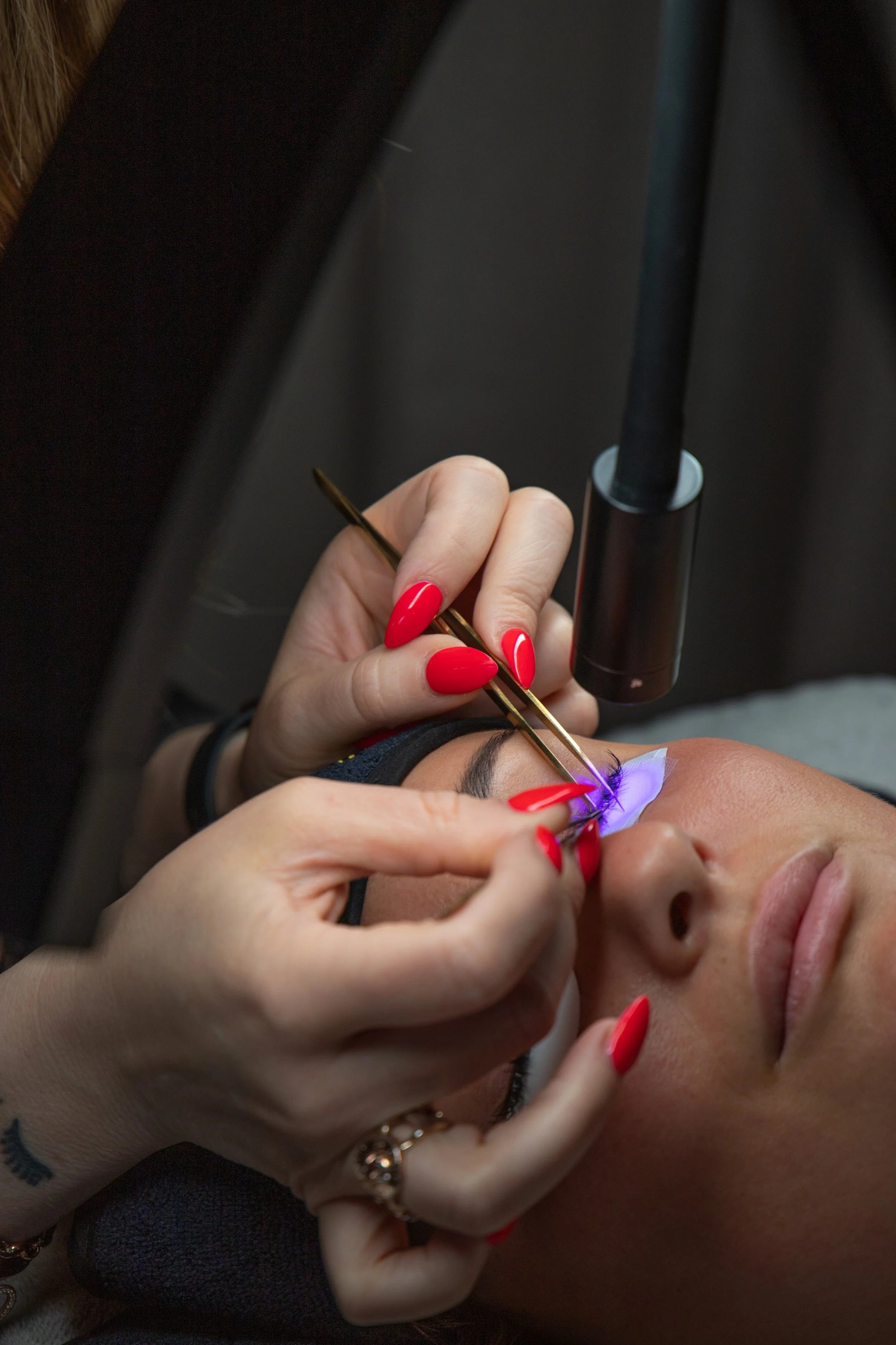 A person receiving a lash extension treatment, lying down with eyes closed, while a beautician applies extensions with tweezers under a UV light.