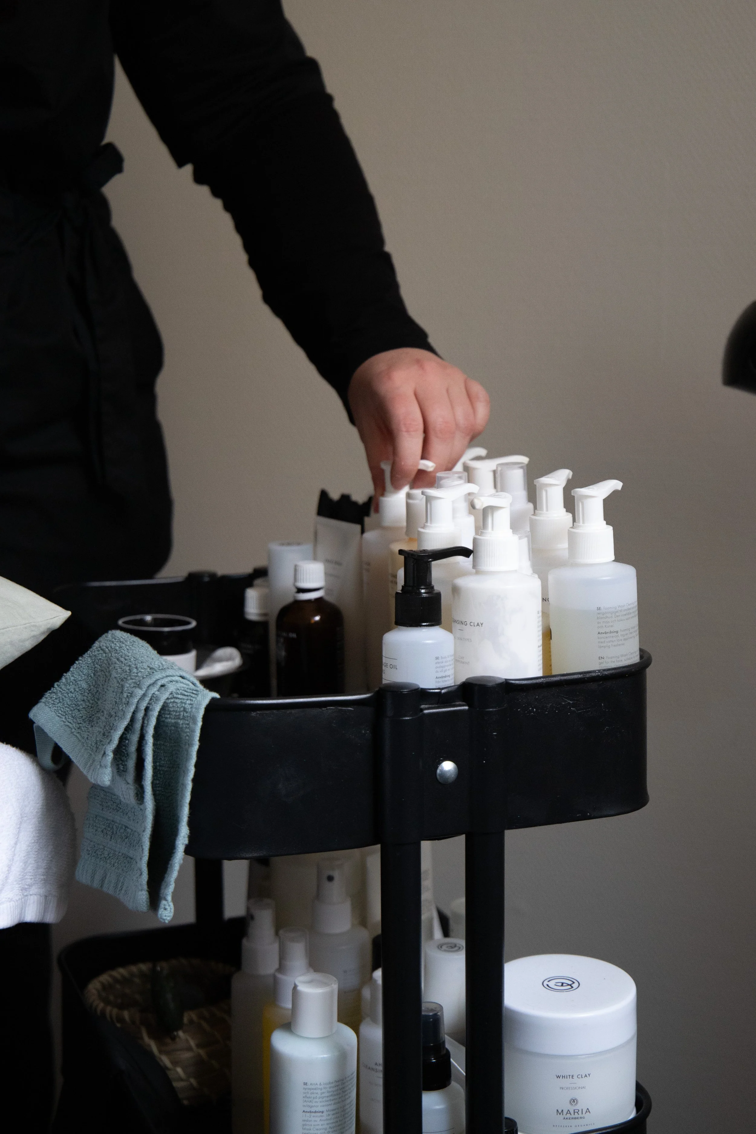 A person in black clothing reaching for bottles of skincare or beauty products on a black cart, with towels hanging over the side and a woven basket on the bottom shelf.