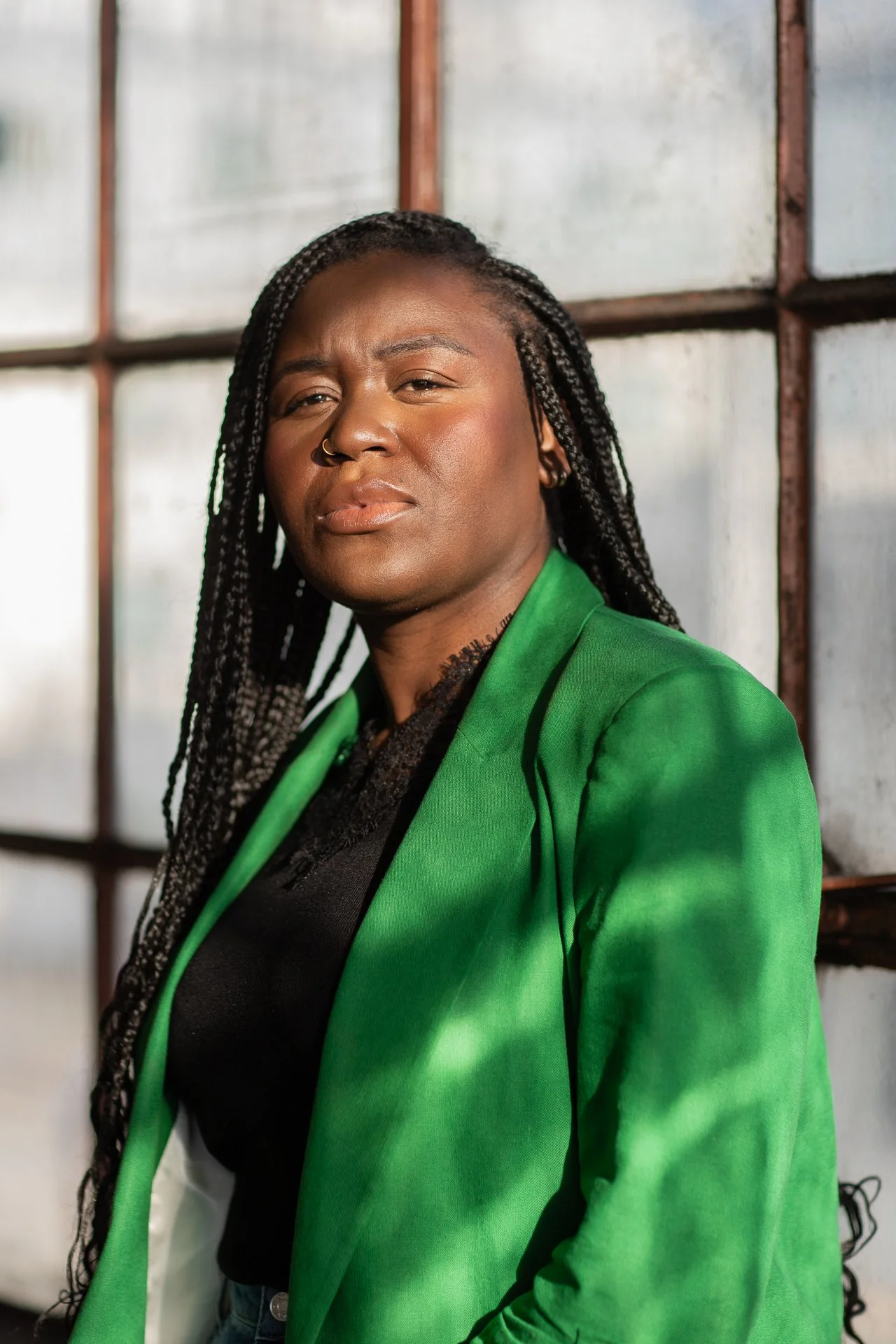 Portrait of a woman with braided hair, wearing a green blazer and black top, standing in front of a large industrial window.