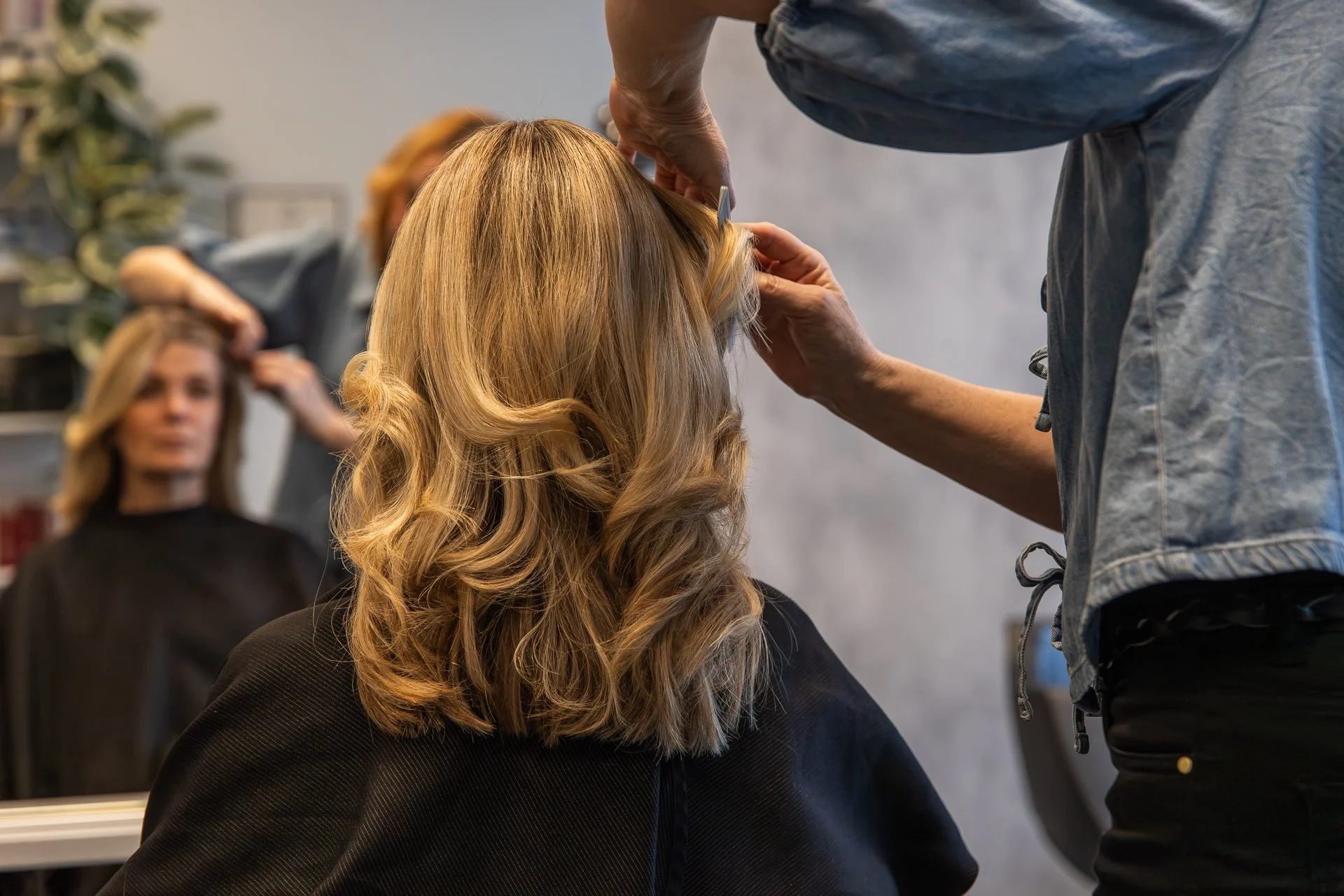 A hairdresser styling a woman's blonde, curled hair in a salon with a mirror and other clients visible in the background.