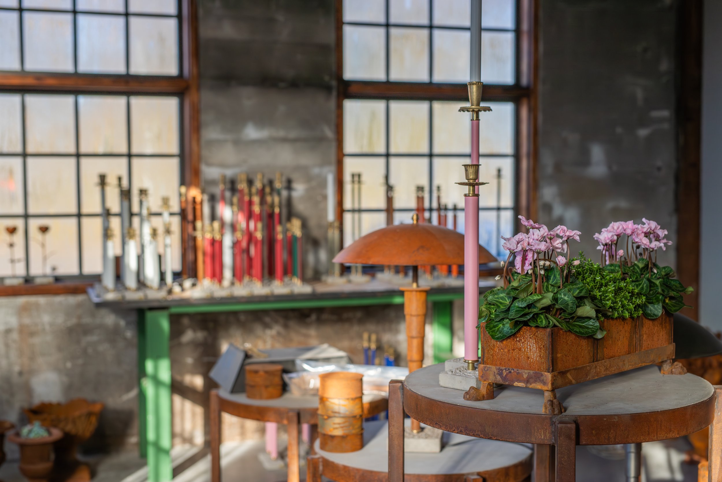 Interior of an art studio or workshop with a rustic brick wall, large grid windows, and a variety of candles, flowers, and craft supplies on tables.