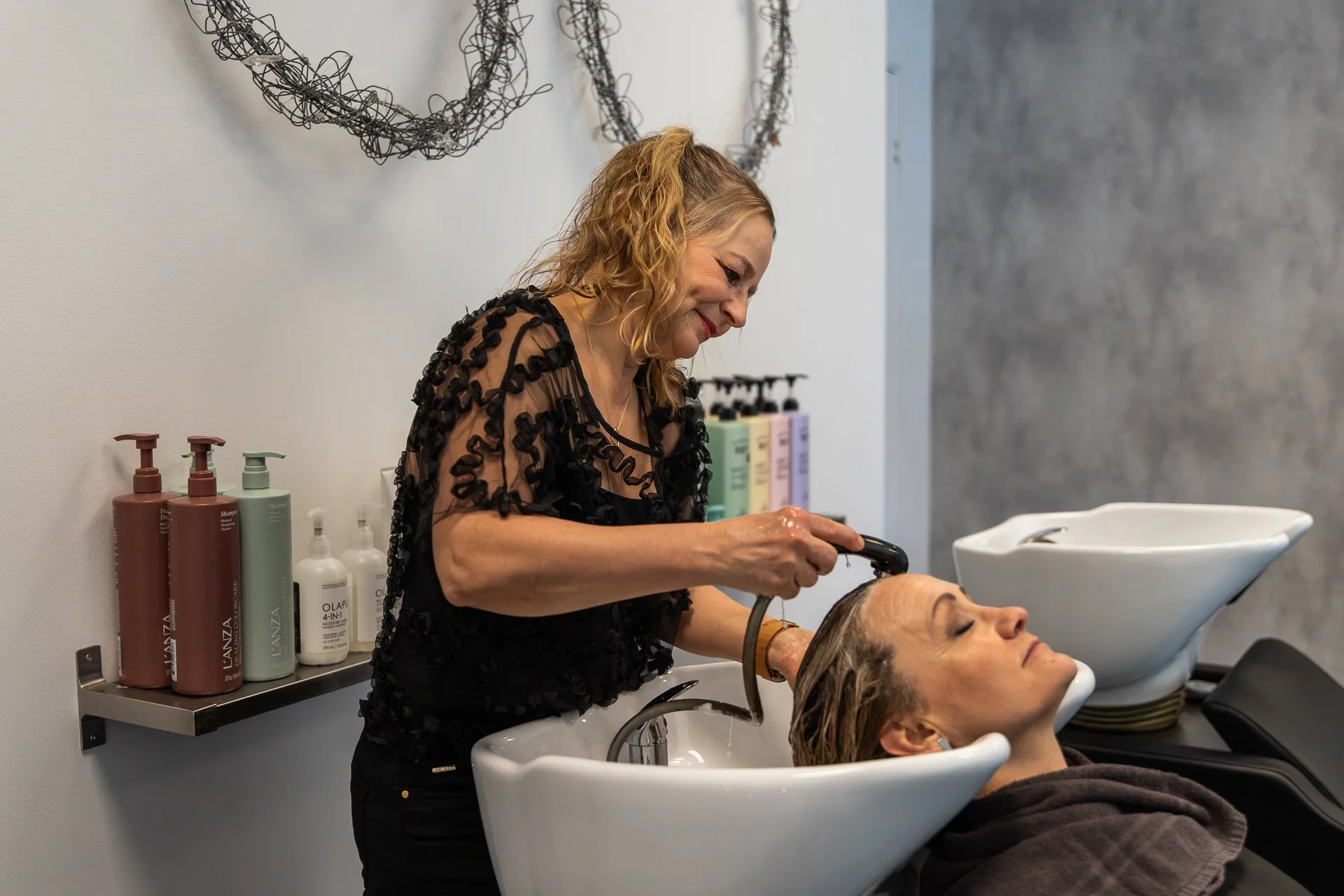 A woman is washing a man's hair in a salon while the man reclines with his eyes closed. The woman smiling looks focused on her task. Behind her, there is a shelf with various hair care products and decorative wire art on the wall.