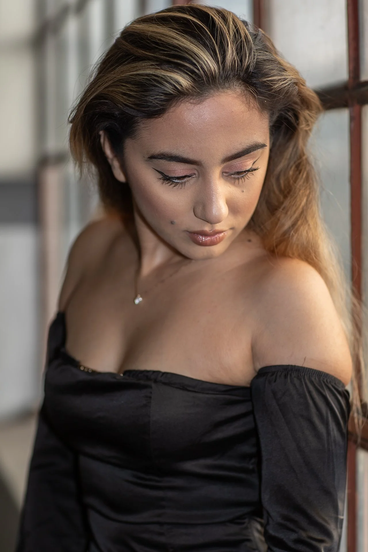 A woman with wavy, blonde-brown hair looking down, wearing a black off-the-shoulder top and a delicate necklace with a small pendant.