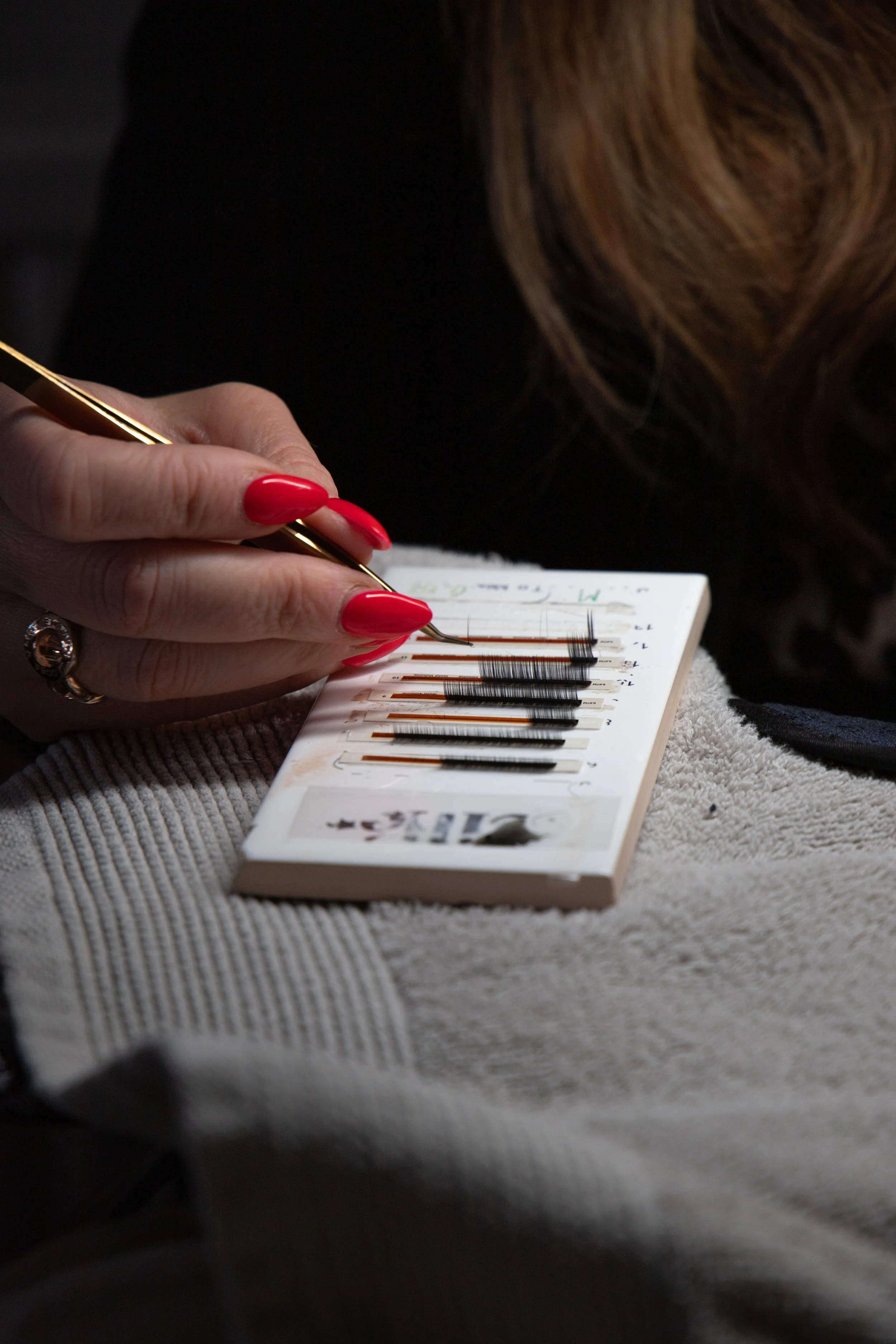 A person with red nail polish using a specialized tool to apply eyelash extensions to a model. The person is holding the tool over a sheet with multiple eyelash extension samples.