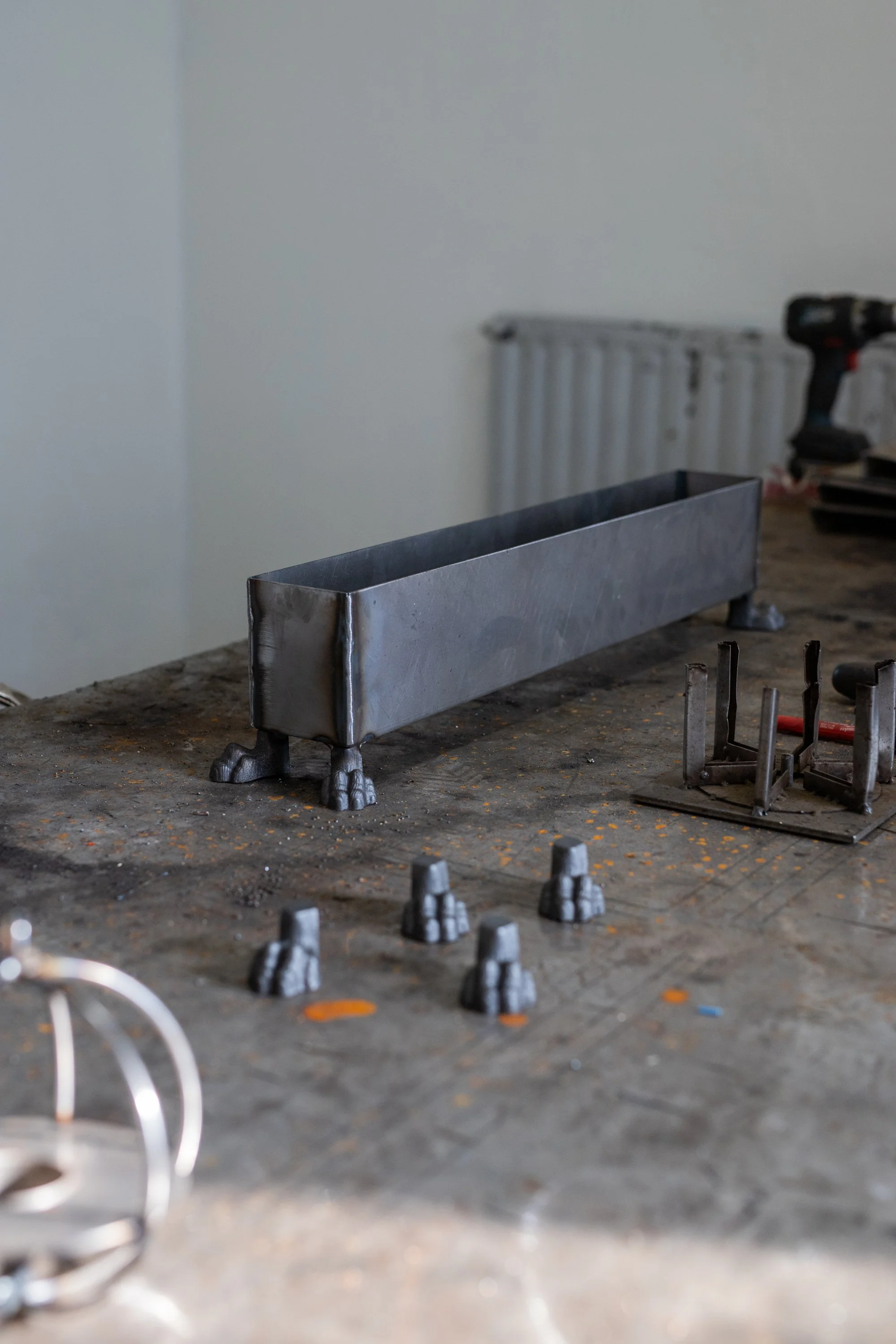 A metal workpiece with clawed feet on a workshop table, surrounded by small metal parts, tools, and a cordless drill in the background.