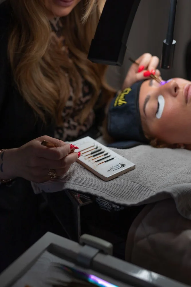 A woman lying down receiving eyelash extensions, with a technician applying the extensions using tweezers and a laser light, in a salon or spa setting.