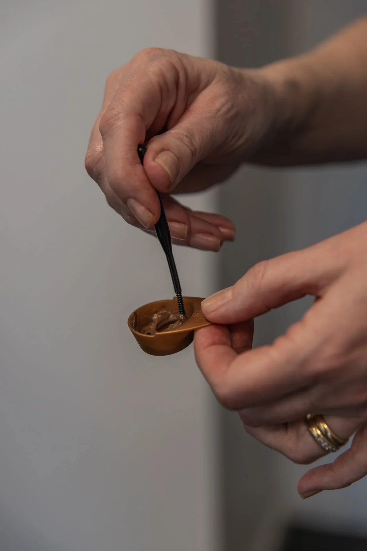 A person is using a small black spoon to scoop a chocolate spread from a small brown bowl.