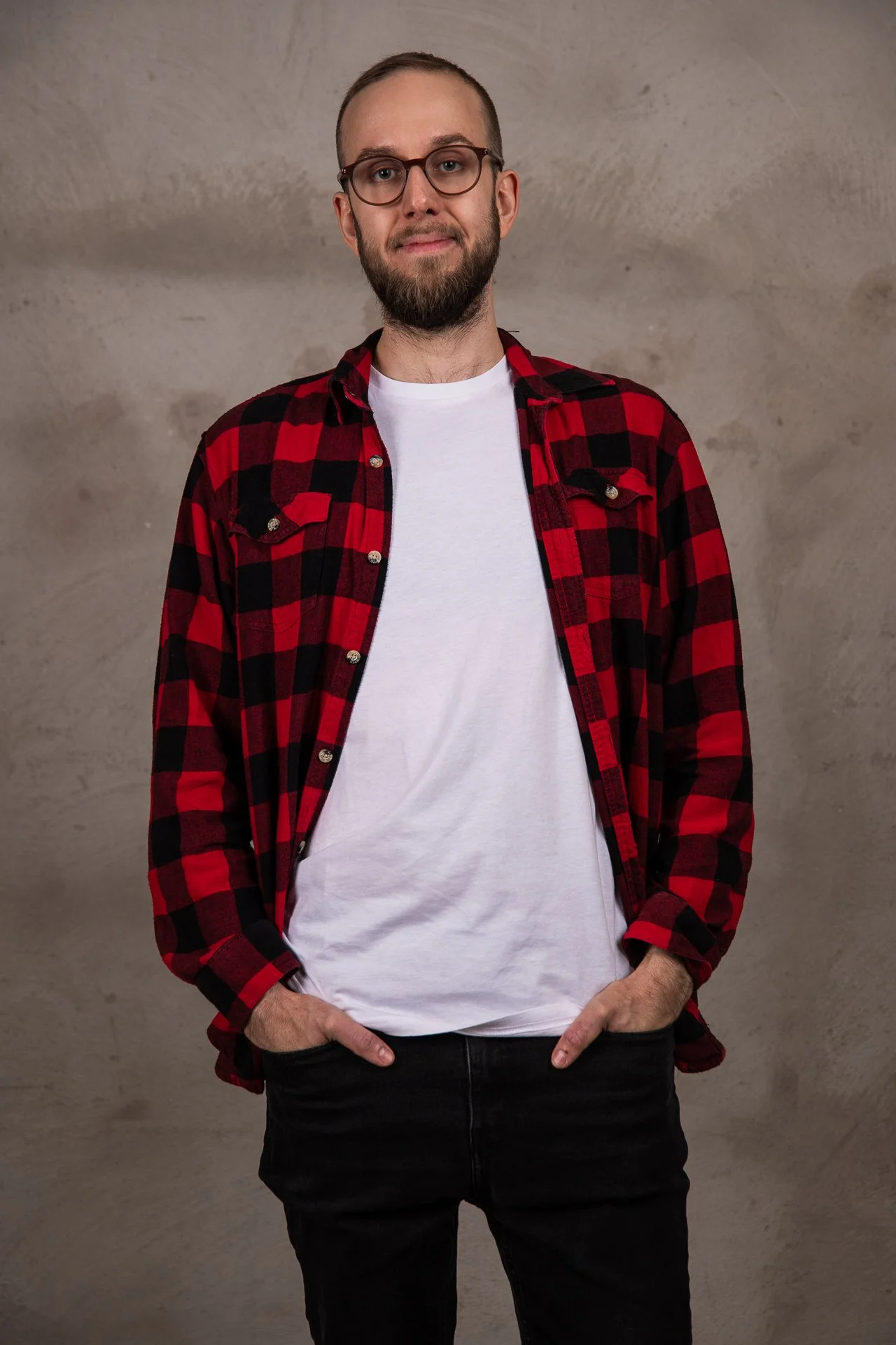 A man with glasses and a beard wearing a red and black plaid shirt over a white t-shirt, standing against a plain, textured wall.