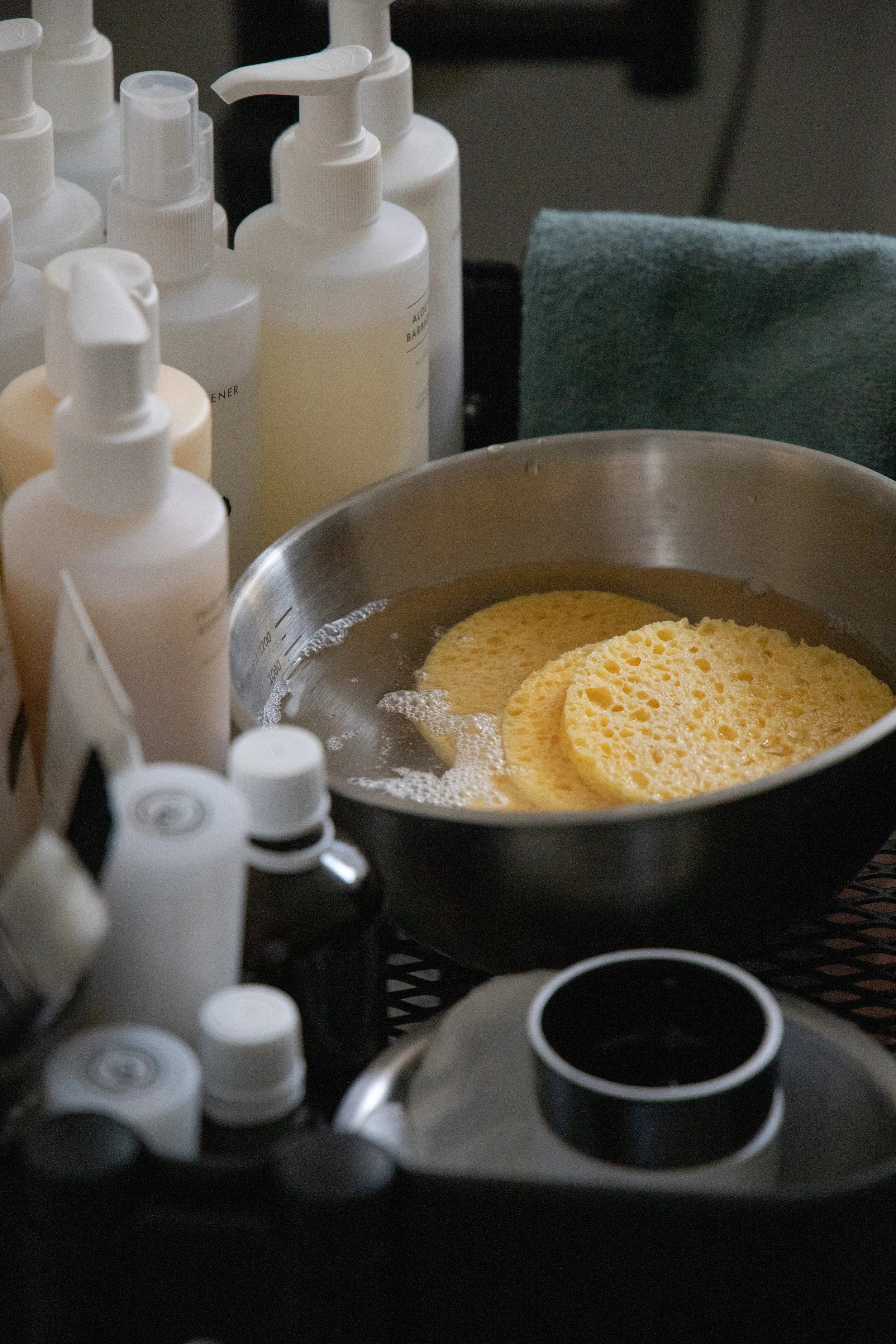 A metal bowl with yellow sponges soaking in water, surrounded by various bottles of skincare or cosmetic products on a countertop, with a green towel and some additional containers nearby.