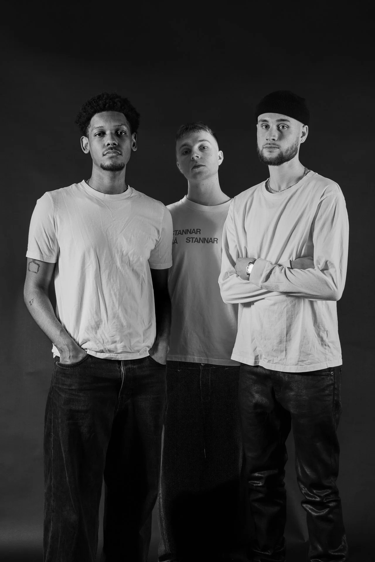Black and white photo of three young men standing against a dark background. They are dressed casually in t-shirts and jeans. The man on the left has an afro hairstyle and tattoos on his left arm. The man in the middle has short hair and is wearing a